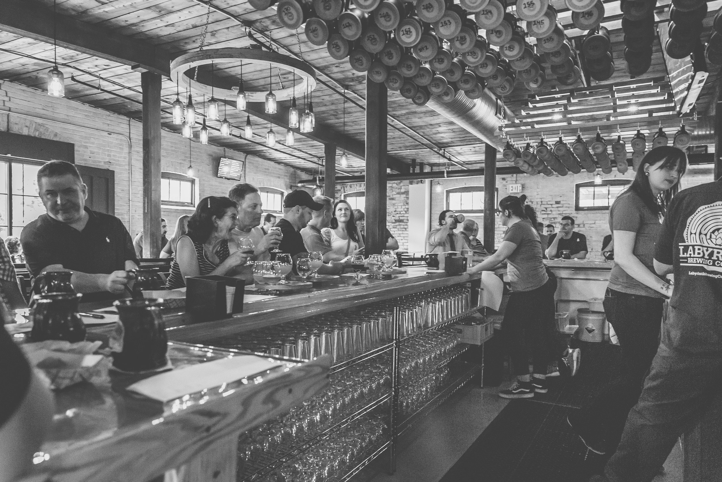 Black and white photo of people sitting at a bar, with drinks and glasses on the counter, and bartenders serving behind the bar in a rustic-style restaurant or brewery.
