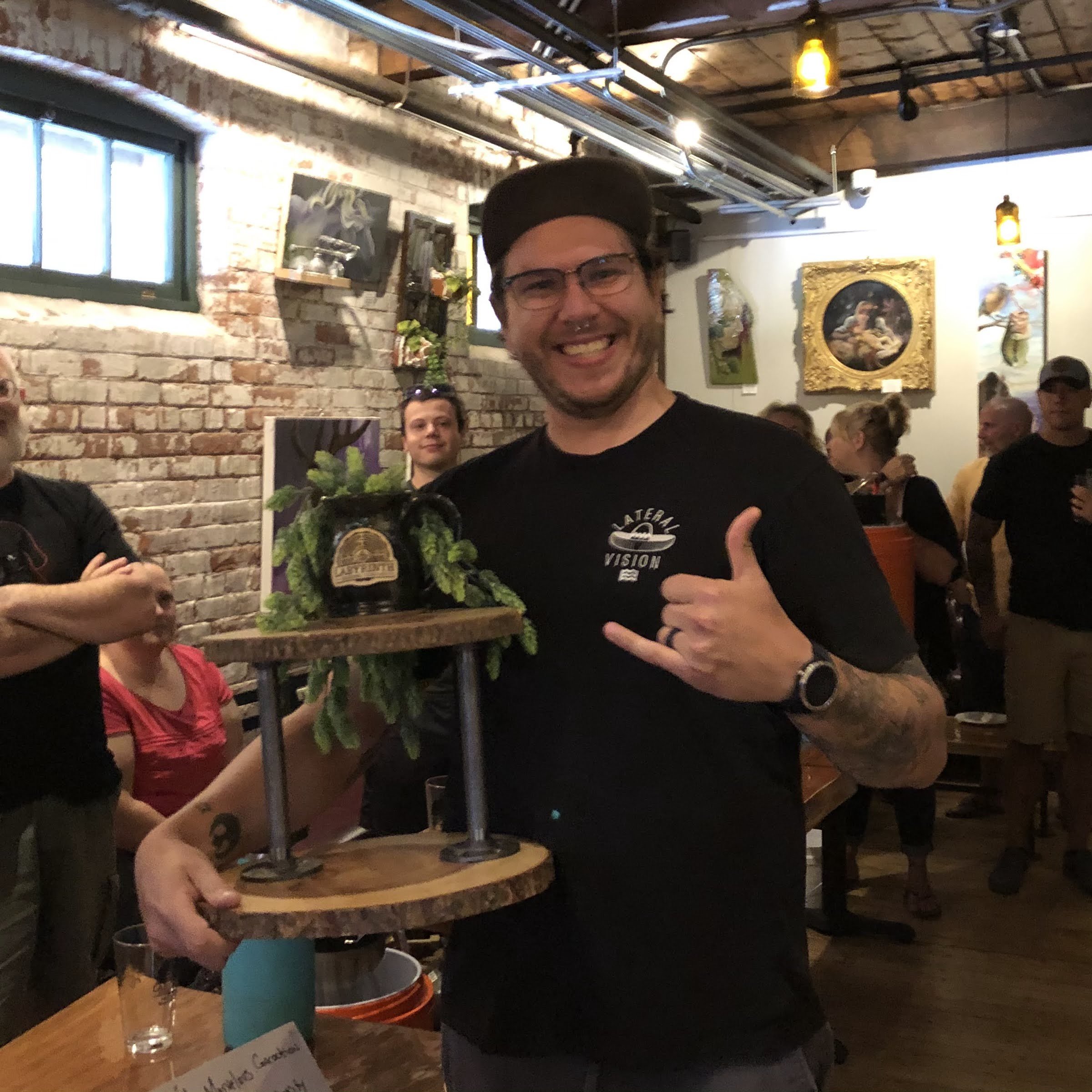 A man smiling and making a shaka sign while holding a wooden stand with a beer growler and plants on top, inside a brewery or pub with brick walls and artwork.