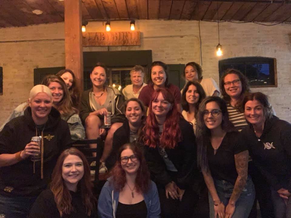Group of women and girls gathered together in a cozy indoor space, smiling at the camera, with warm lighting and brick walls in the background.