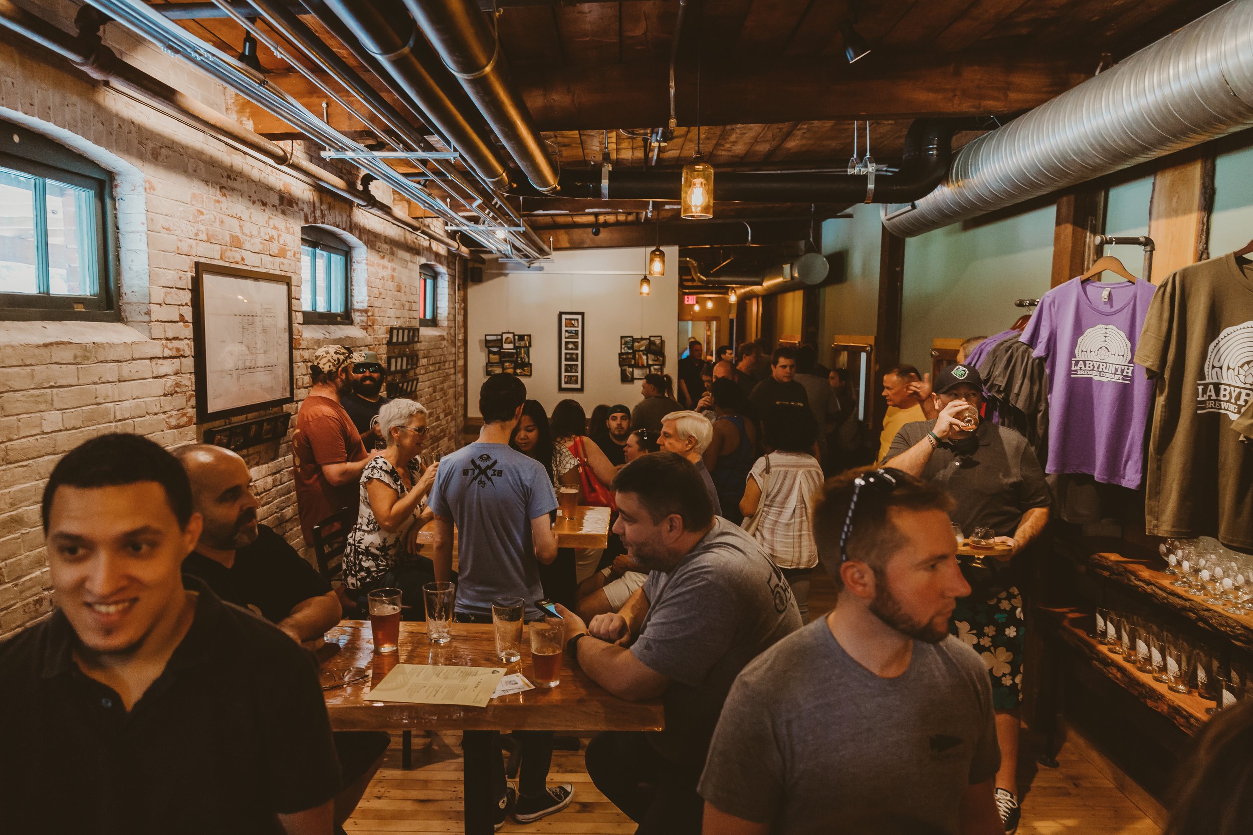 Crowd of people inside a brewery with exposed brick walls, industrial ceiling piping, and merchandise on display.