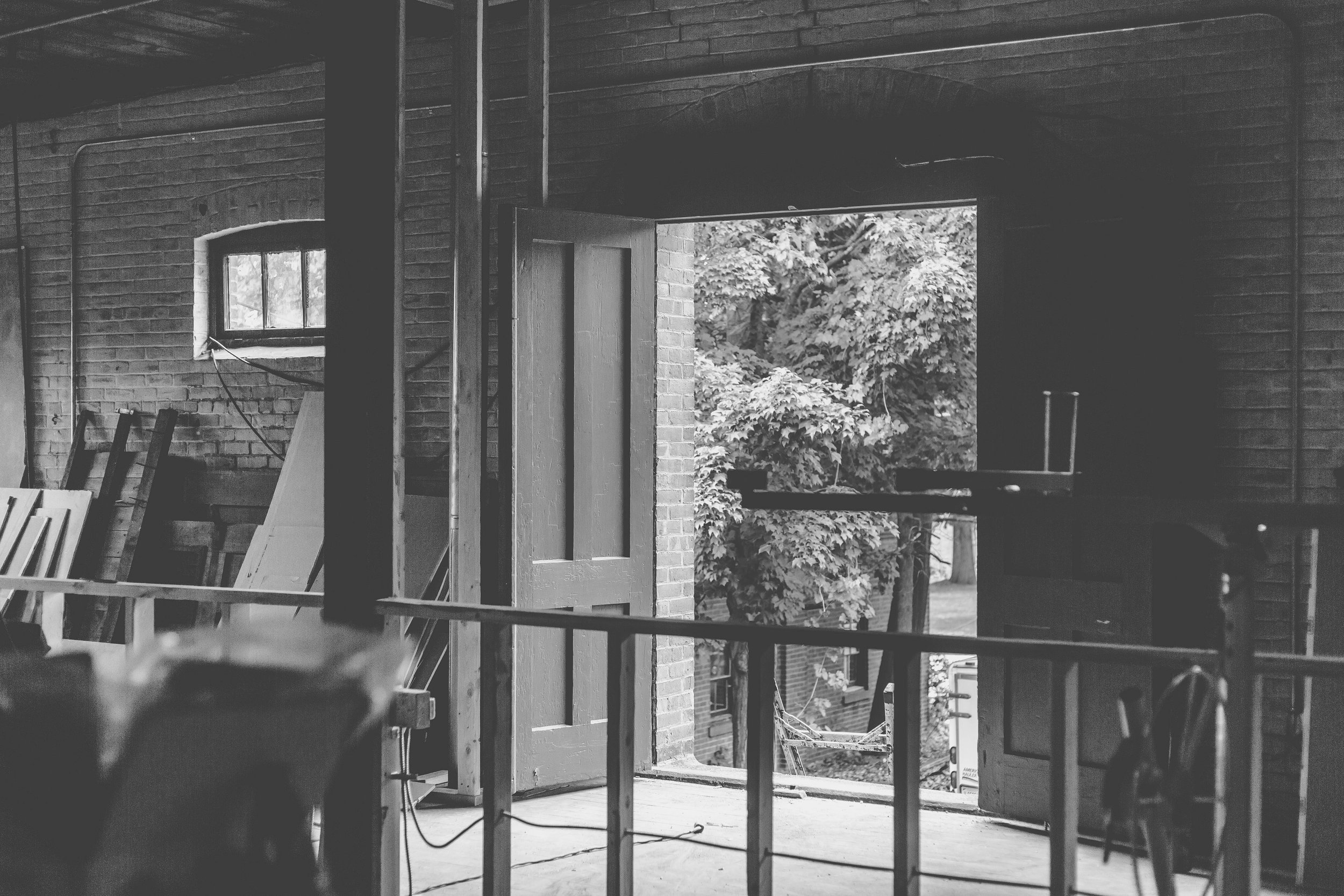 Interior view of a building under construction or renovation, with an open doorway, exposed brick walls, wood framing, and construction materials, overlooking trees outside.