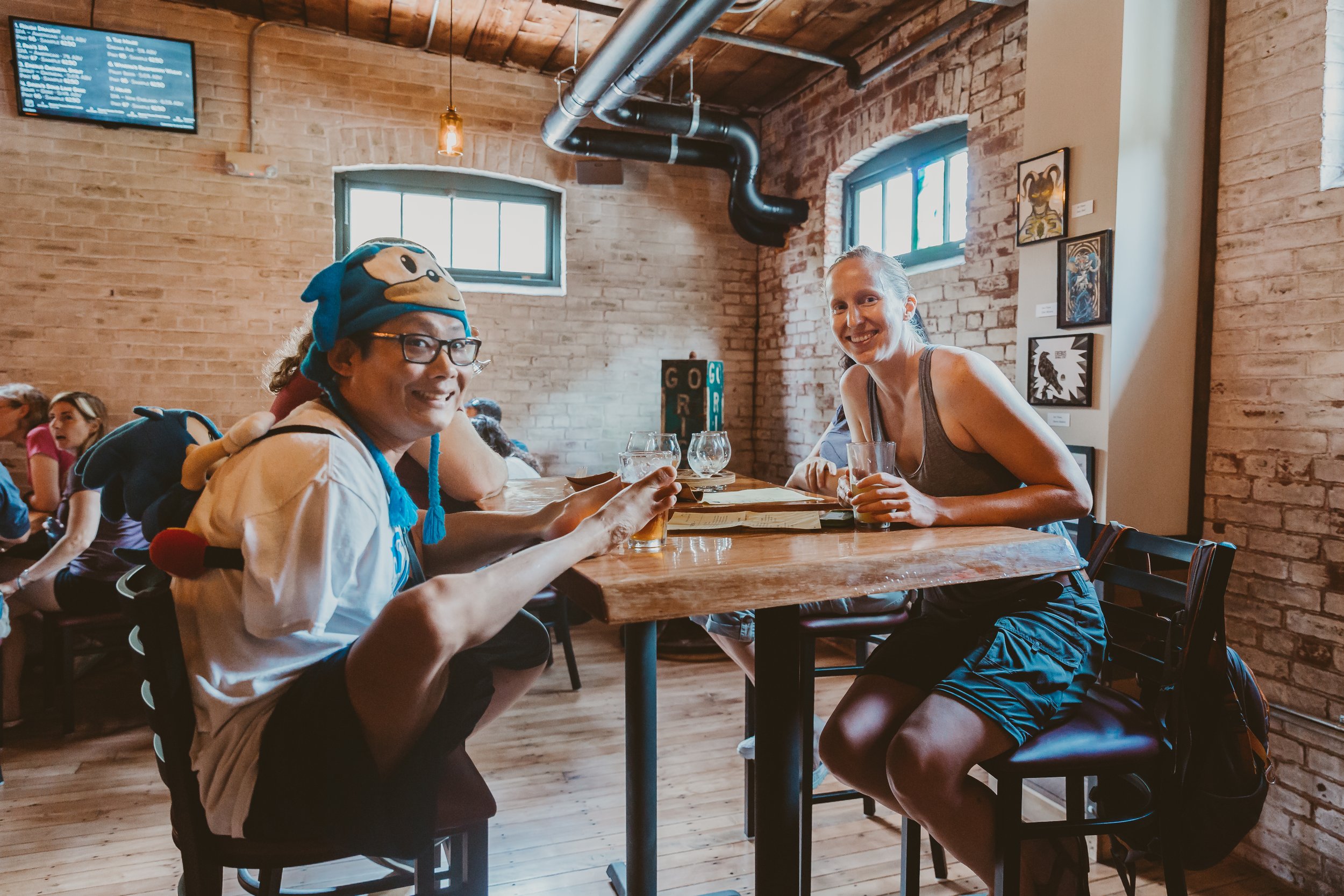 Two women sitting at a wooden table in a brick-walled cafe, smiling and raising their glasses for a toast. A man wearing a Sonic the Hedgehog hat is partially visible in the background.