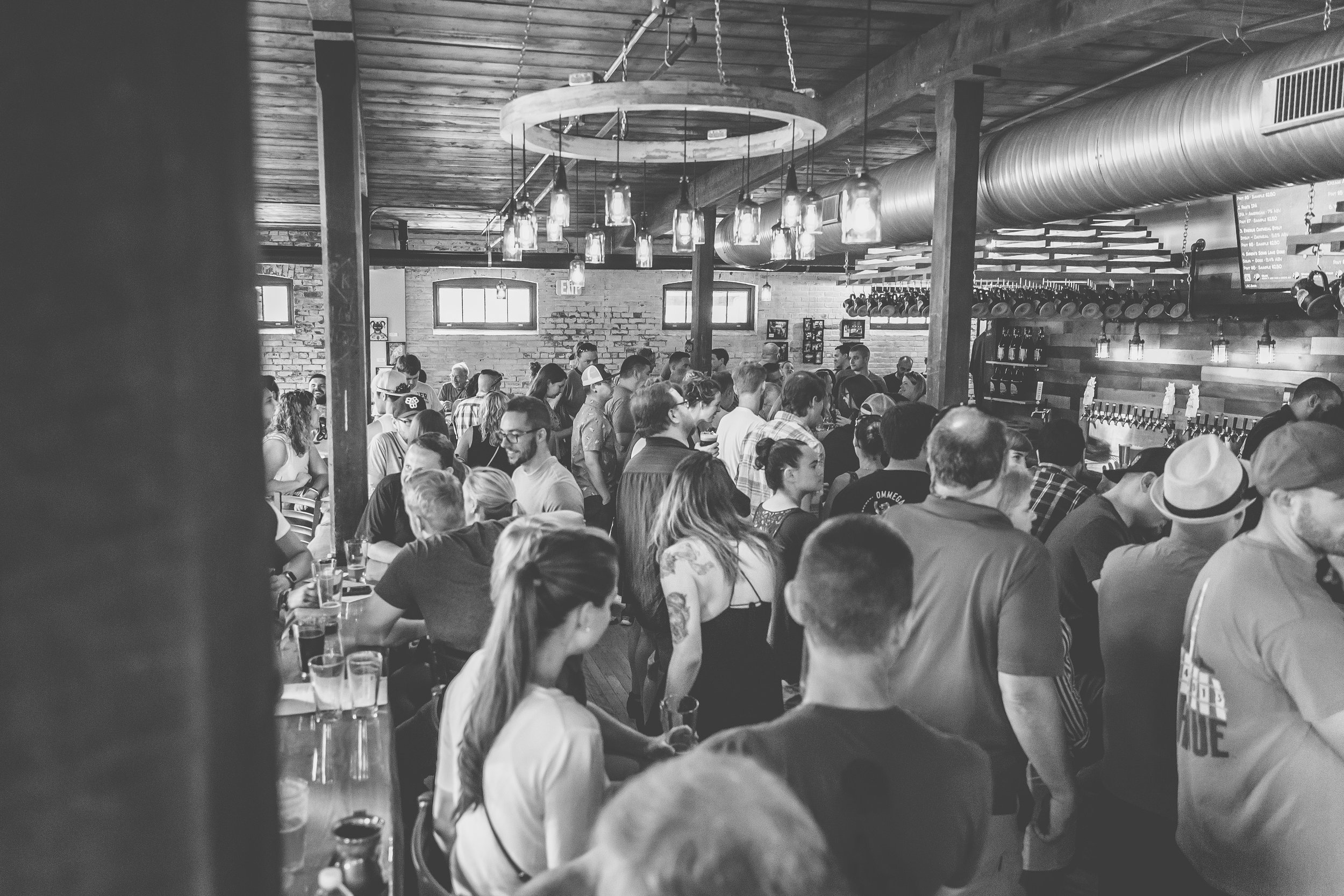 Crowded bar or restaurant with people standing, sitting, and socializing inside, decorated with warm lighting and rustic wooden decor.