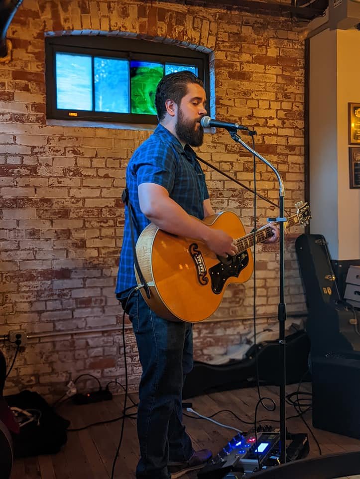 Man with a beard and plaid shirt playing an acoustic guitar and singing into a microphone on a stage with a brick wall and a stained glass window in the background.