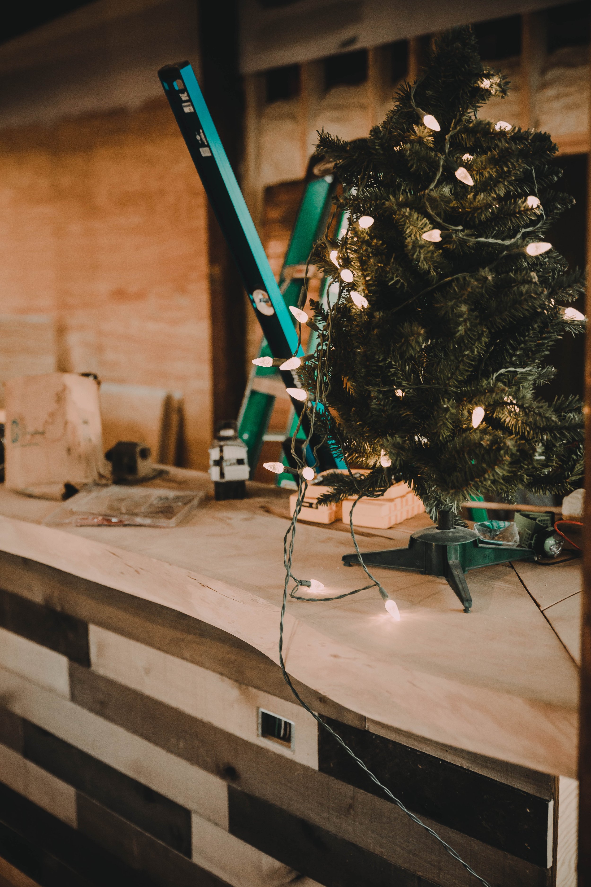 Small decorated Christmas tree with string lights on a wooden table with tools and a work ladder nearby, in a workshop setting.