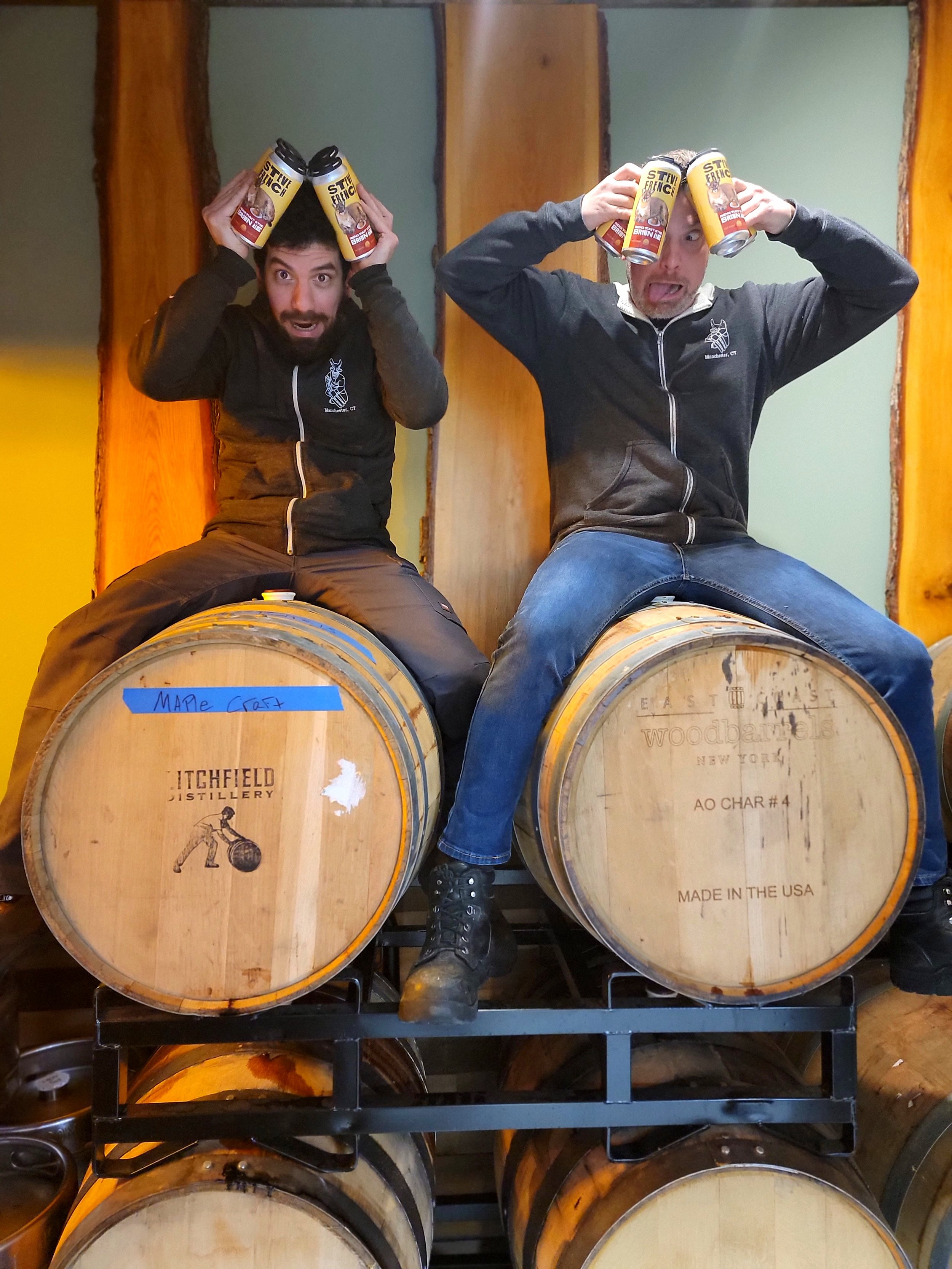 Two men sitting on barrels, holding cans of beer on their heads, making funny faces in a brewery. One man has dark hair and a beard, the other is bald.