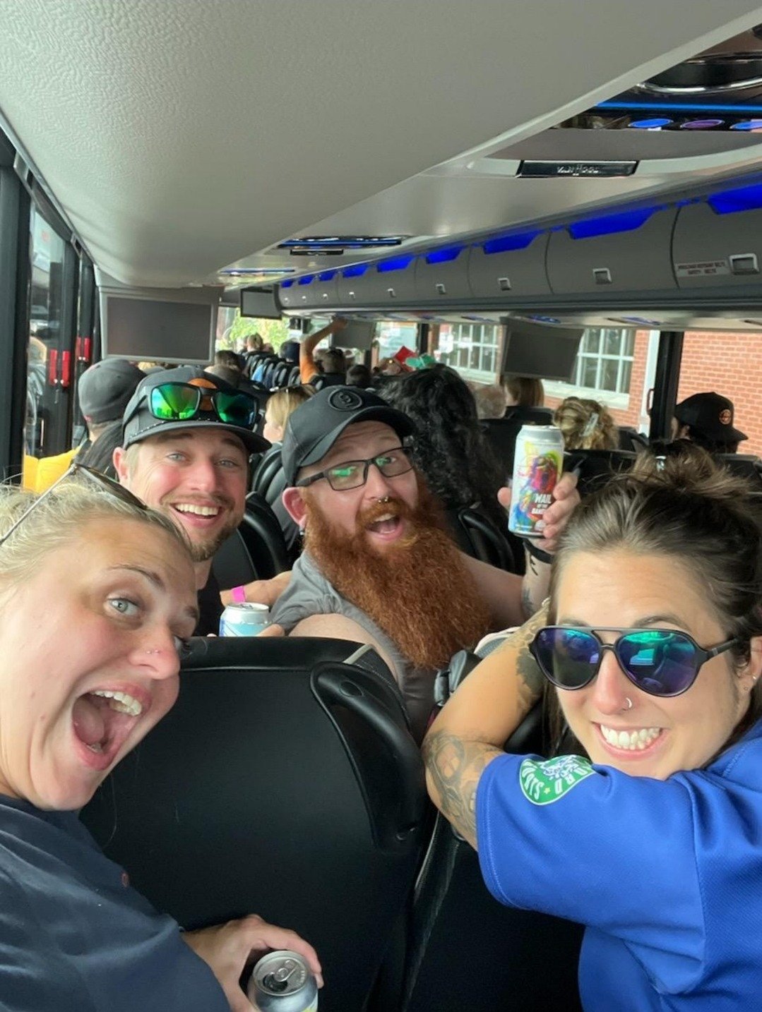 Group of young adults smiling and posing for a photo inside a bus, holding canned drinks, with some wearing sunglasses.