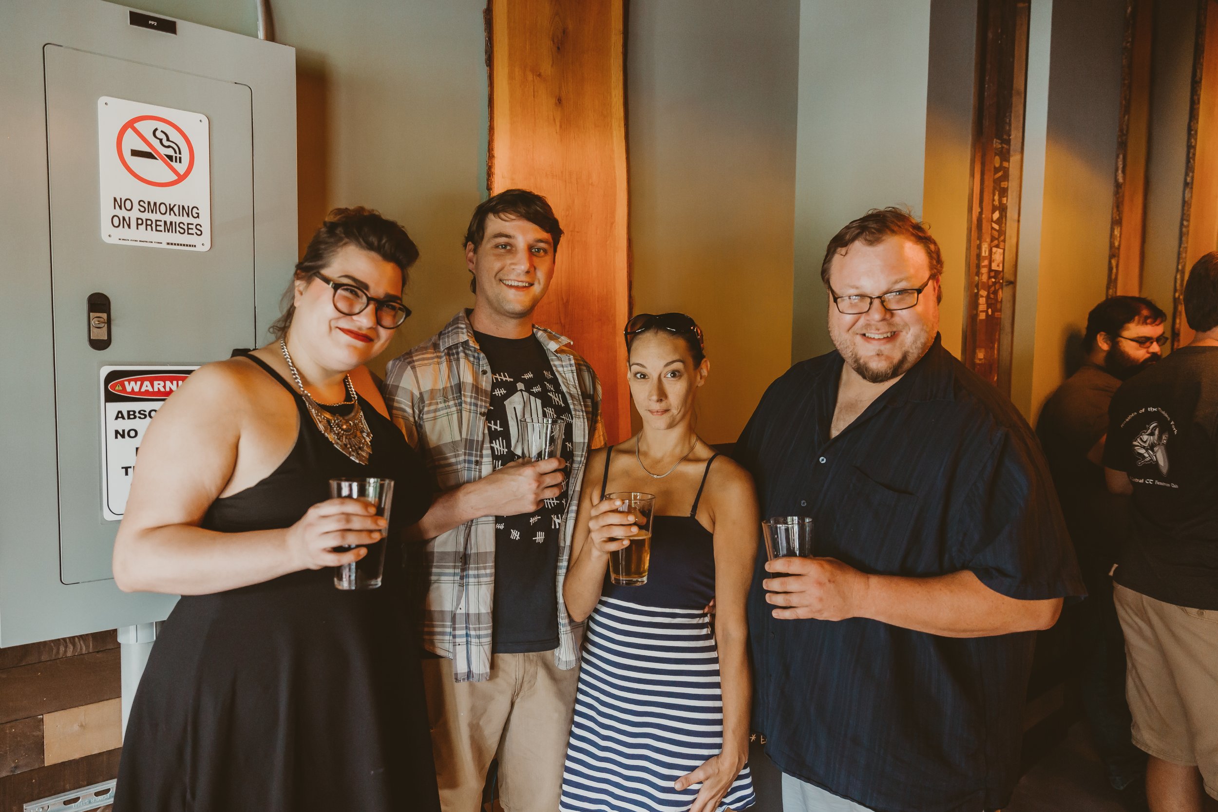 Four people standing together at a social event, holding glasses of drinks and smiling, in an indoor setting.