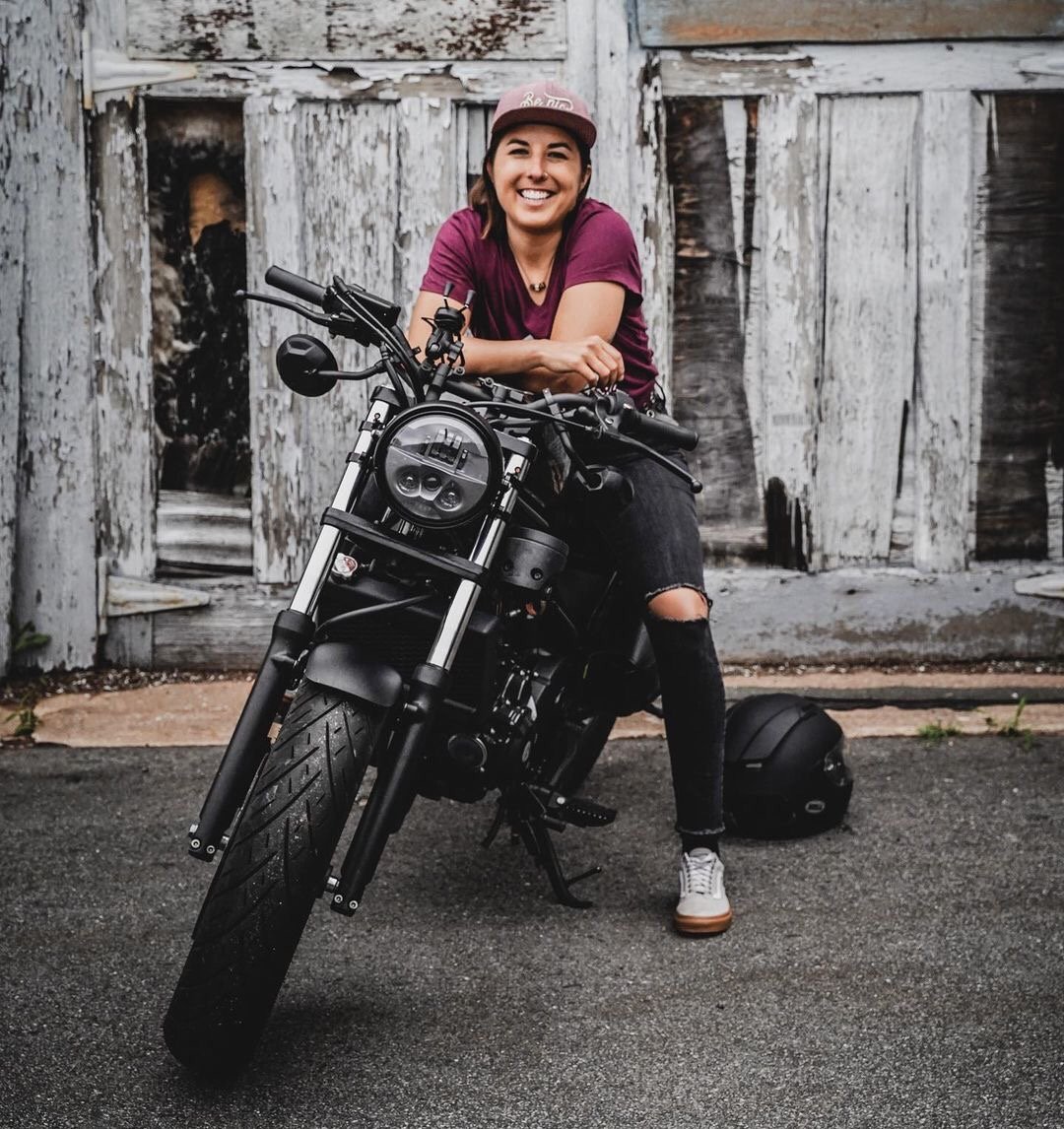 A woman with a beaming smile, wearing a pink cap, maroon T-shirt, ripped black jeans, and white sneakers, leaning on a black motorcycle with a wooden barn in the background.