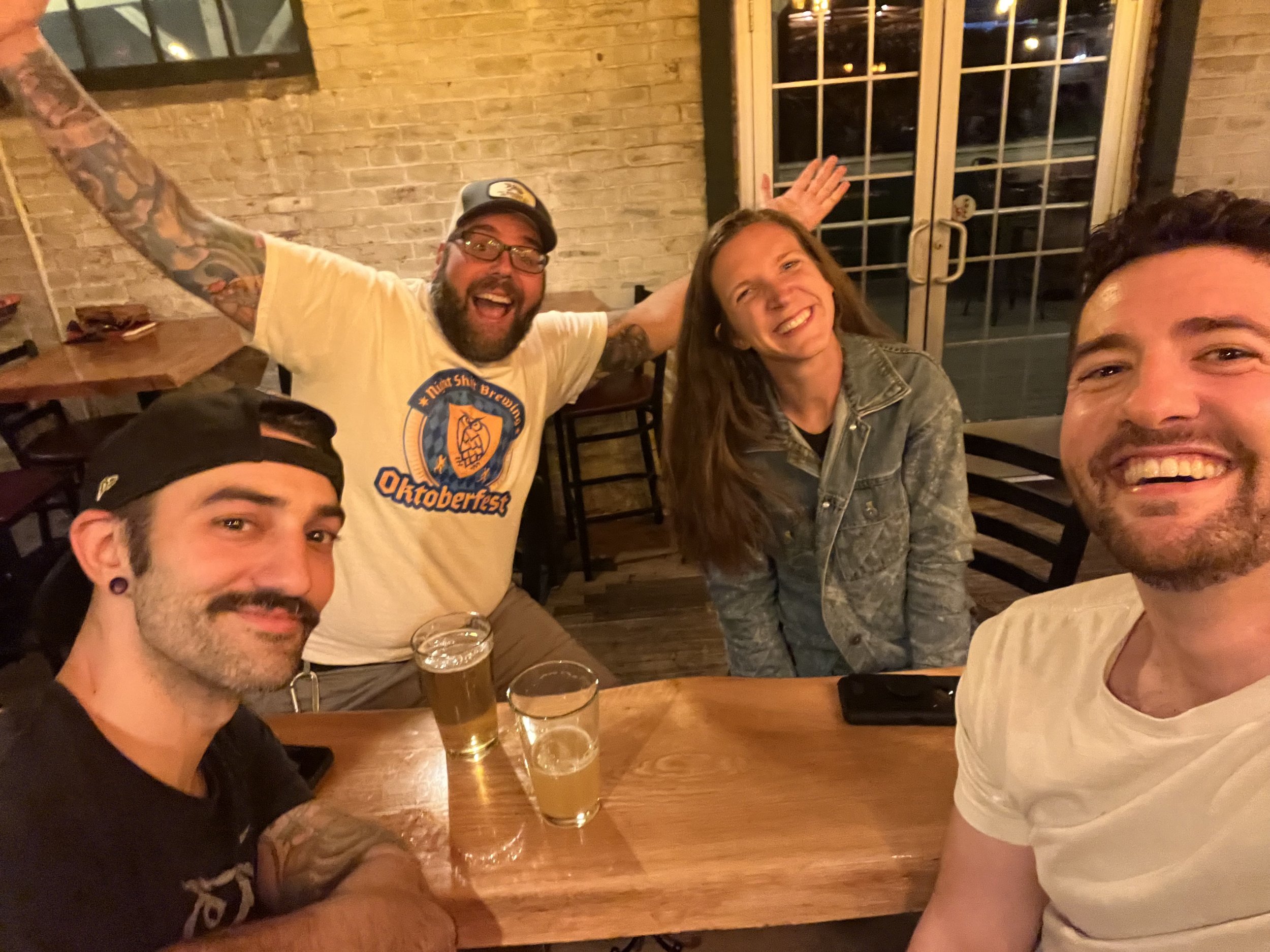 Four friends smiling and celebrating at a wooden table in a cozy bar or restaurant, with glasses of beer and a brick wall in the background.