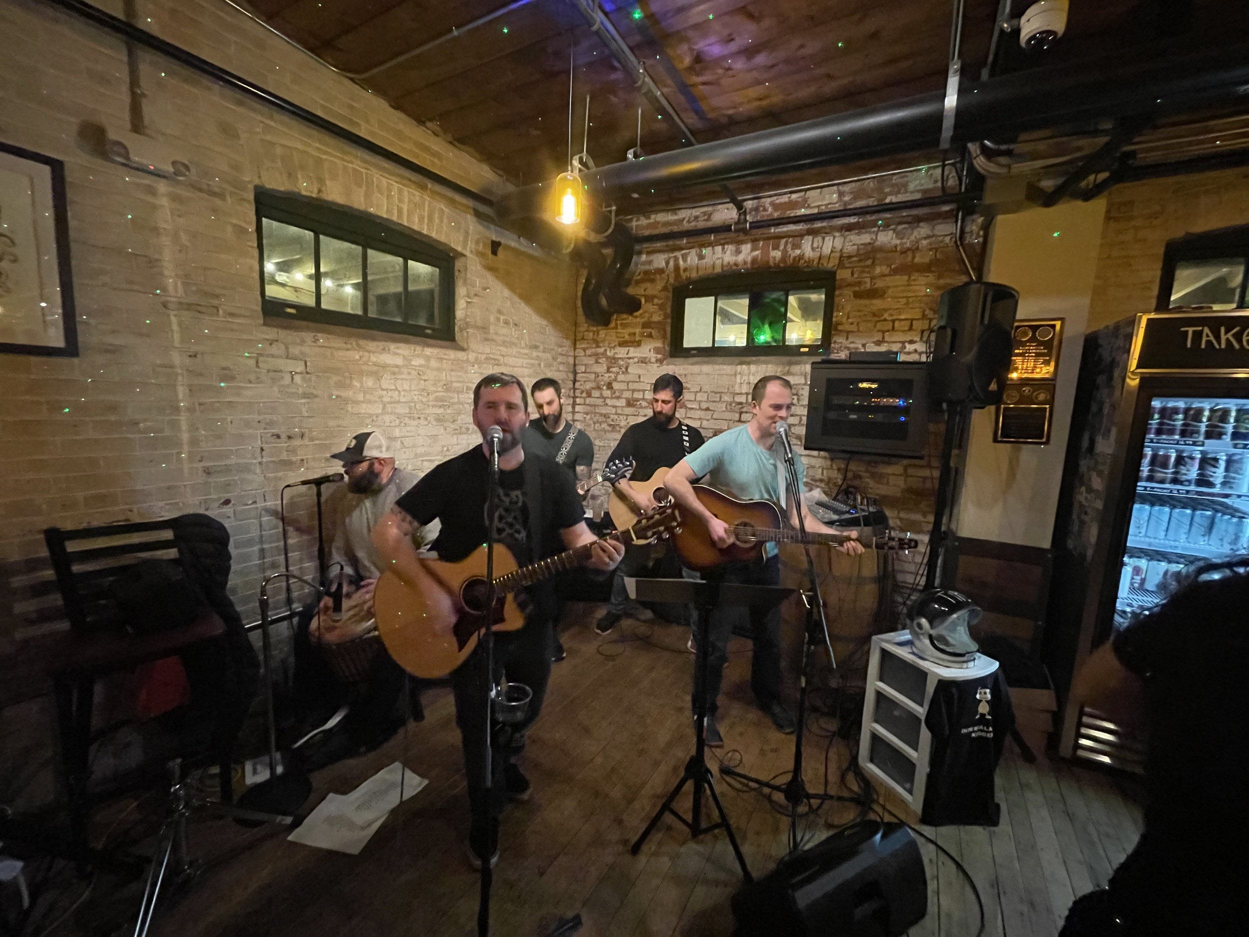 A group of musicians performing live with guitars in a cozy, brick-walled bar or cafe, under warm lighting.