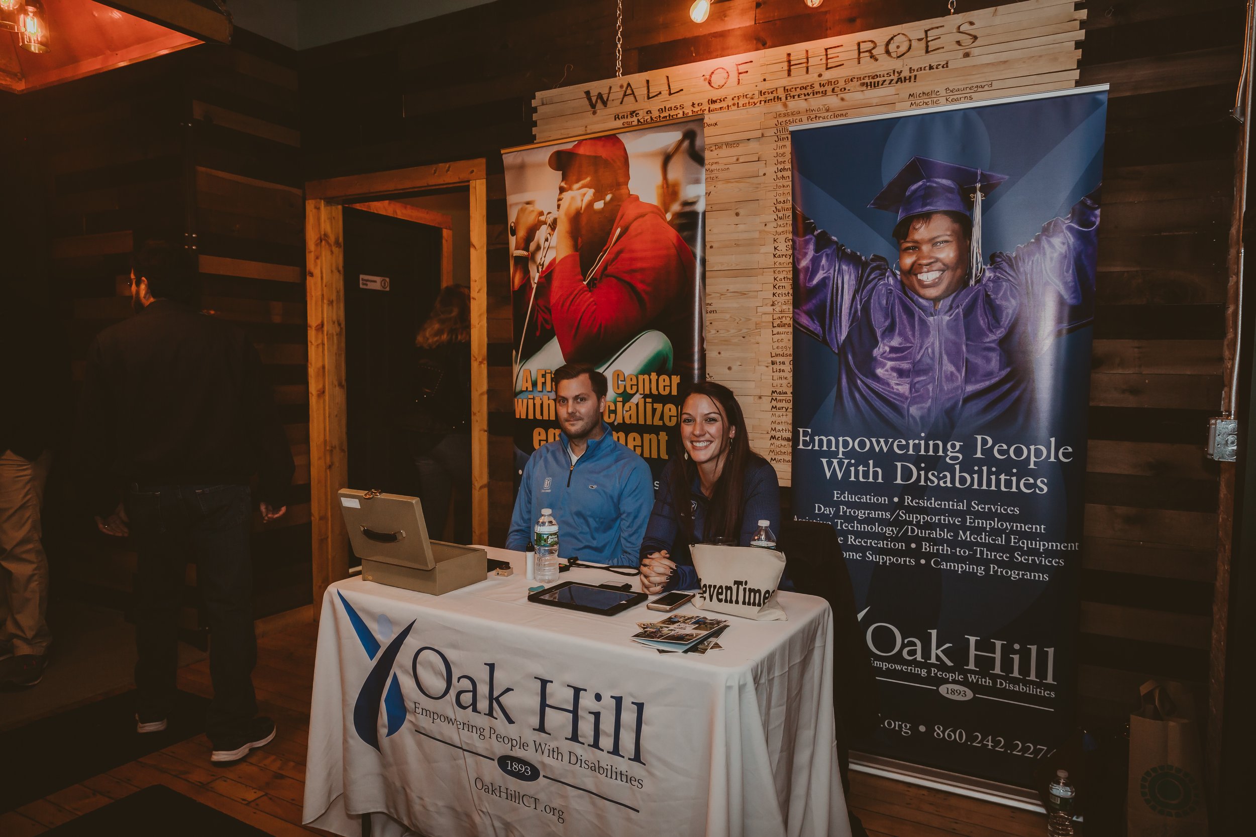 Two people seated at an information booth for Oak Hill, an organization supporting people with disabilities, with banners behind them. One banner features a smiling woman in a graduation cap and gown. The table is covered with a white cloth displayin