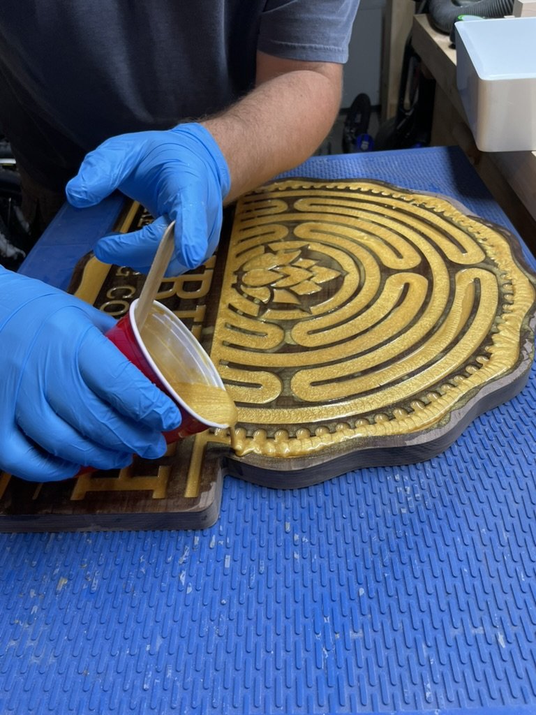 Person in a gray shirt with blue gloves pouring resin into a carved wooden maze puzzle.