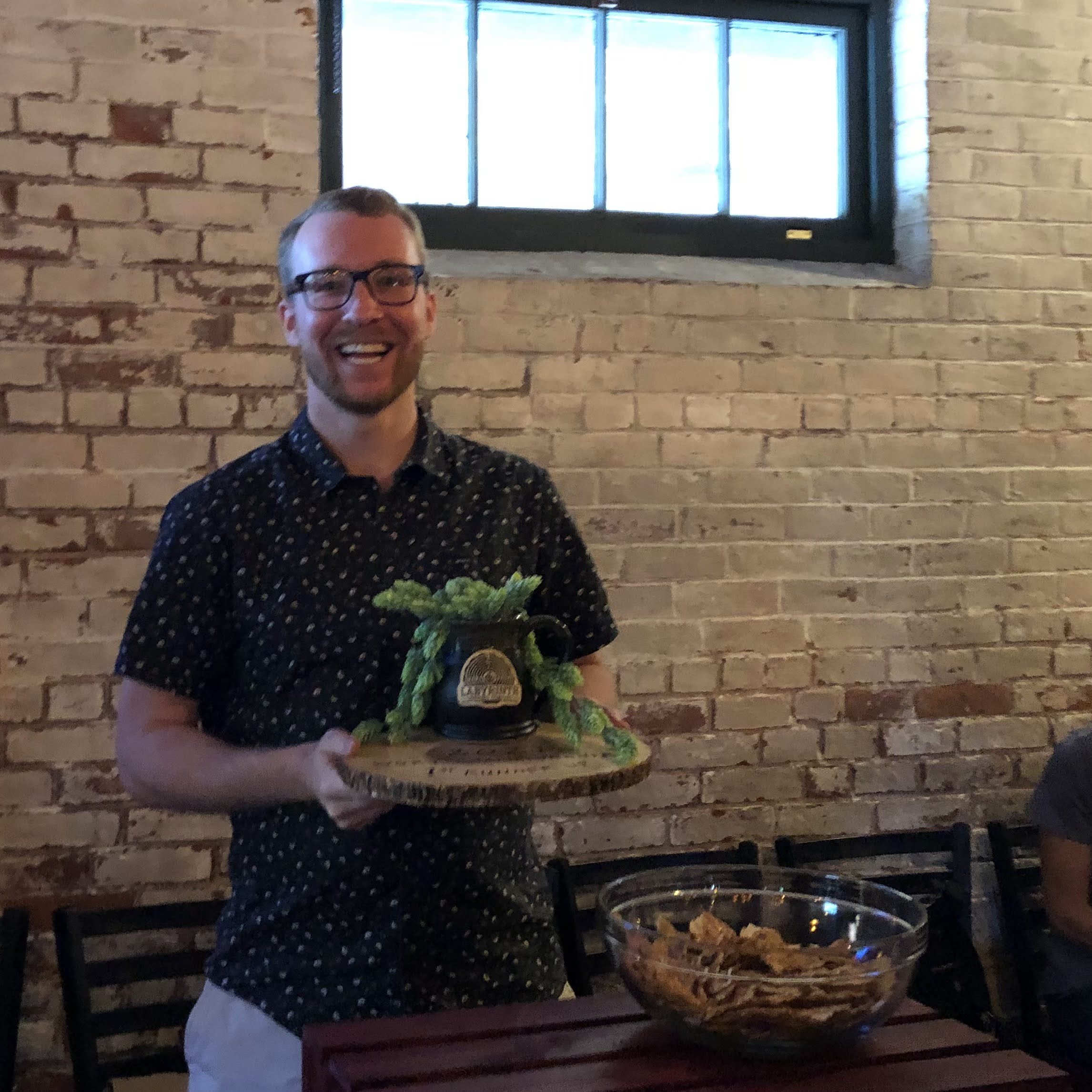 A smiling man wearing glasses holding a wooden tray with a decorative vessel and green foliage, standing in front of a brick wall inside a restaurant or cafe.