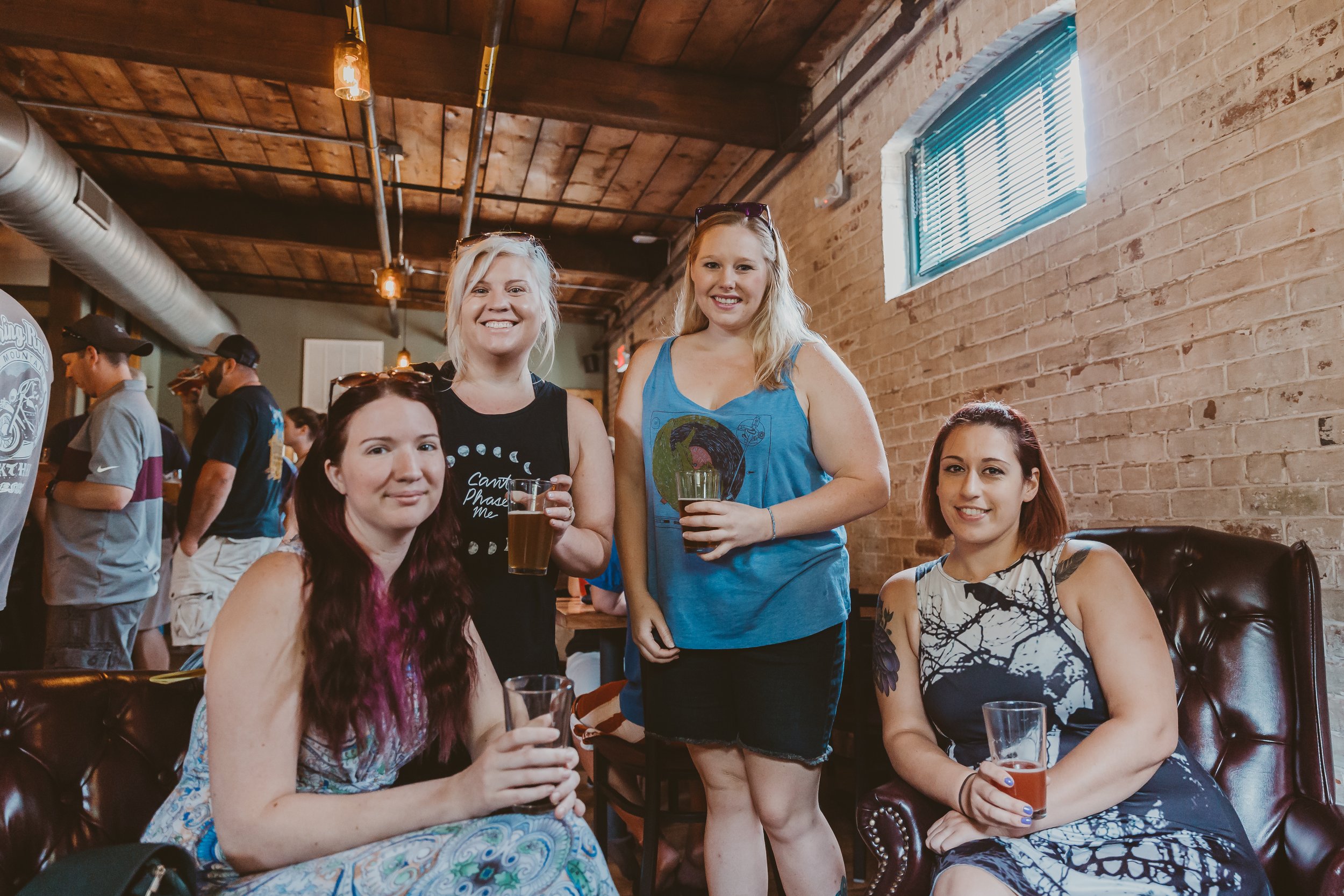 Four women at a bar or pub, smiling and holding drinks, with a brick wall and wooden ceiling in the background.