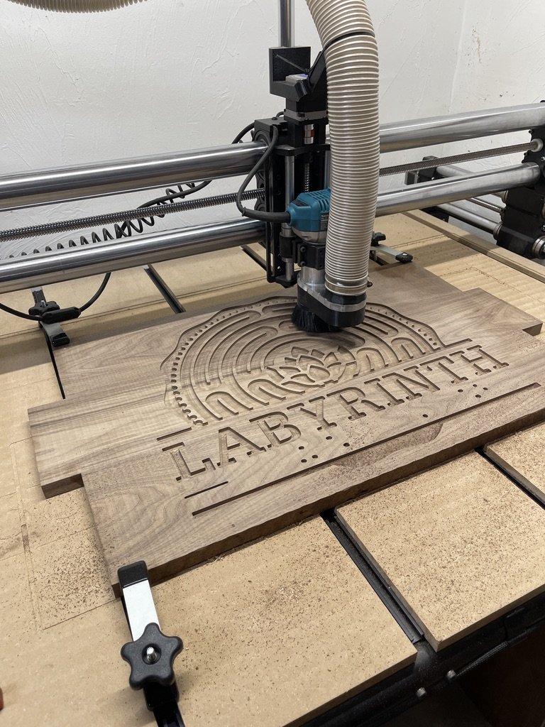 Wooden board with laser engraving of a logo that reads "LABYZRUX" with a circular design and a central image, being engraved by a CNC machine with a dust collection hose attached.