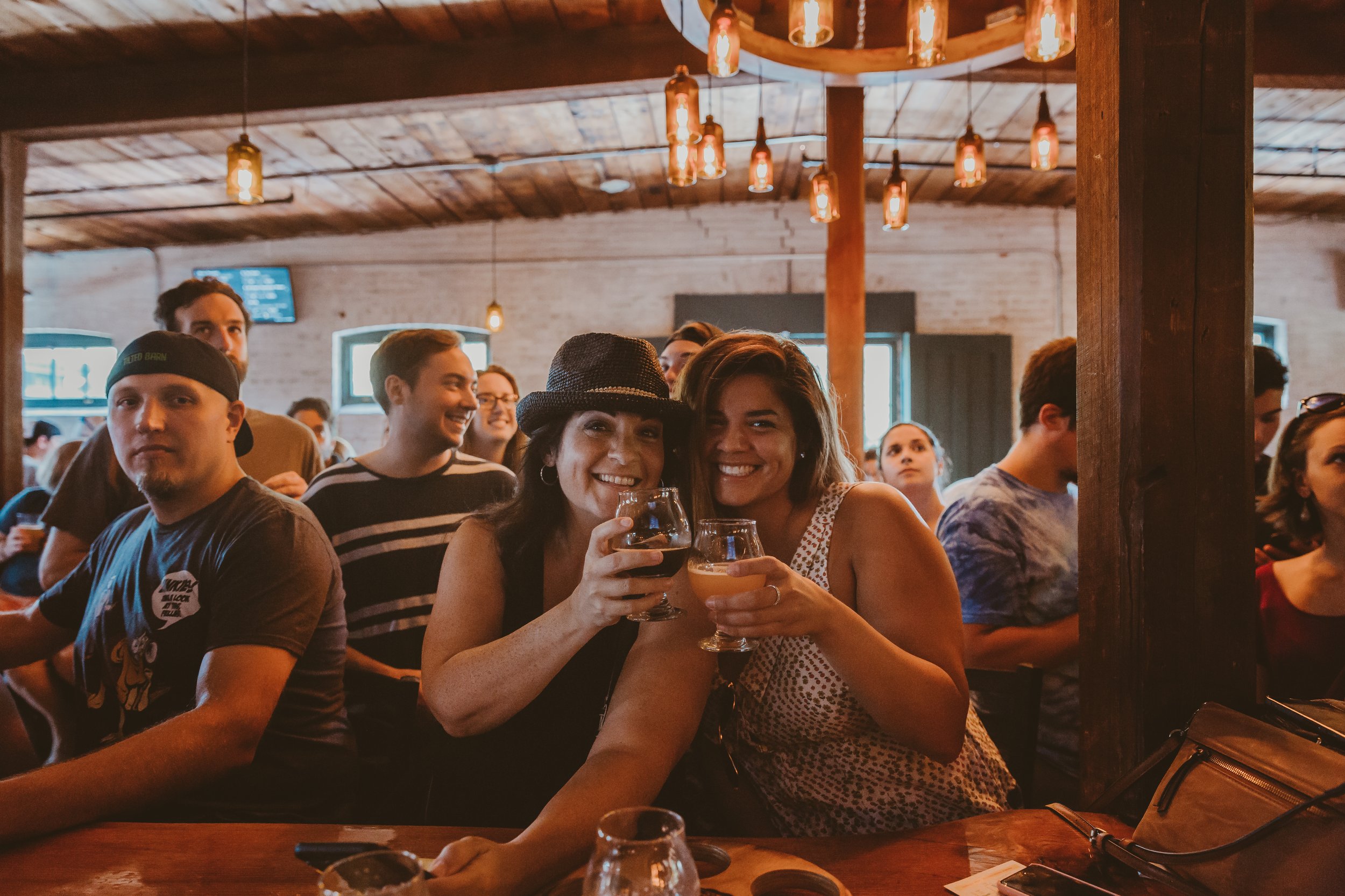 Two women smiling and holding drinks at a bar or restaurant, surrounded by other people, with warm lighting and rustic interior.