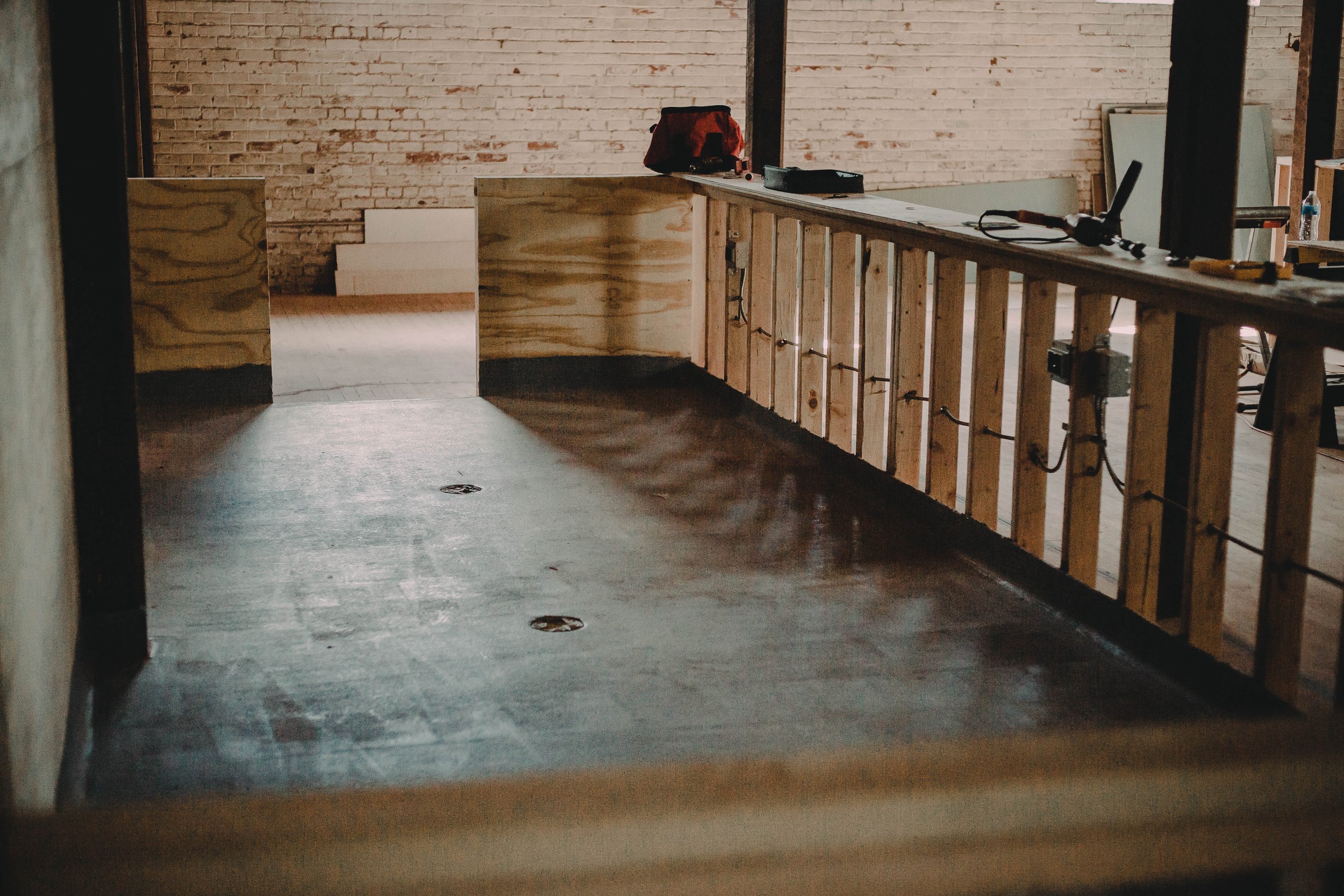 Interior of a building under construction with exposed brick walls, a wooden railing, and a concrete floor with embedded electrical outlets.
