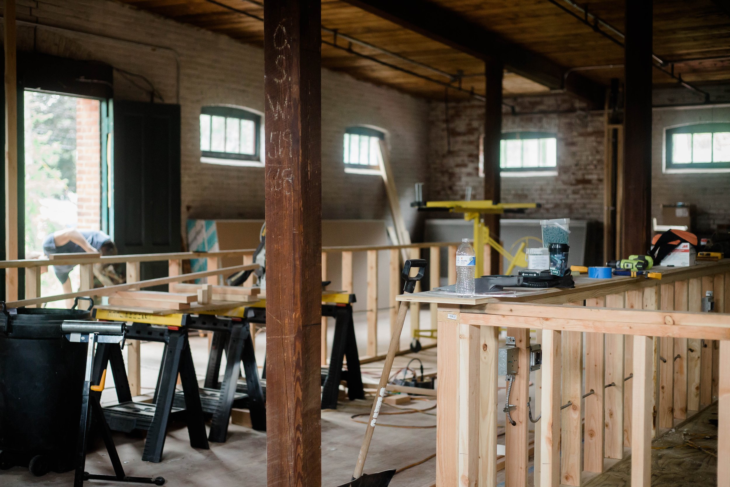 Interior of a building under construction with exposed brick walls, windows, and wooden framing for a kitchen or workspace.
