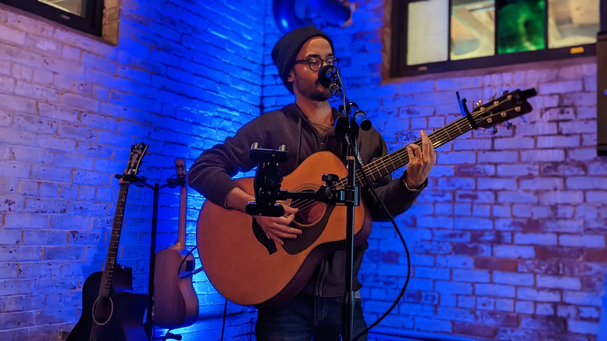 A man wearing a beanie, glasses, and a dark jacket plays an acoustic guitar on stage with a brick wall background illuminated by blue and purple lights. Two additional guitars are on stands behind him.