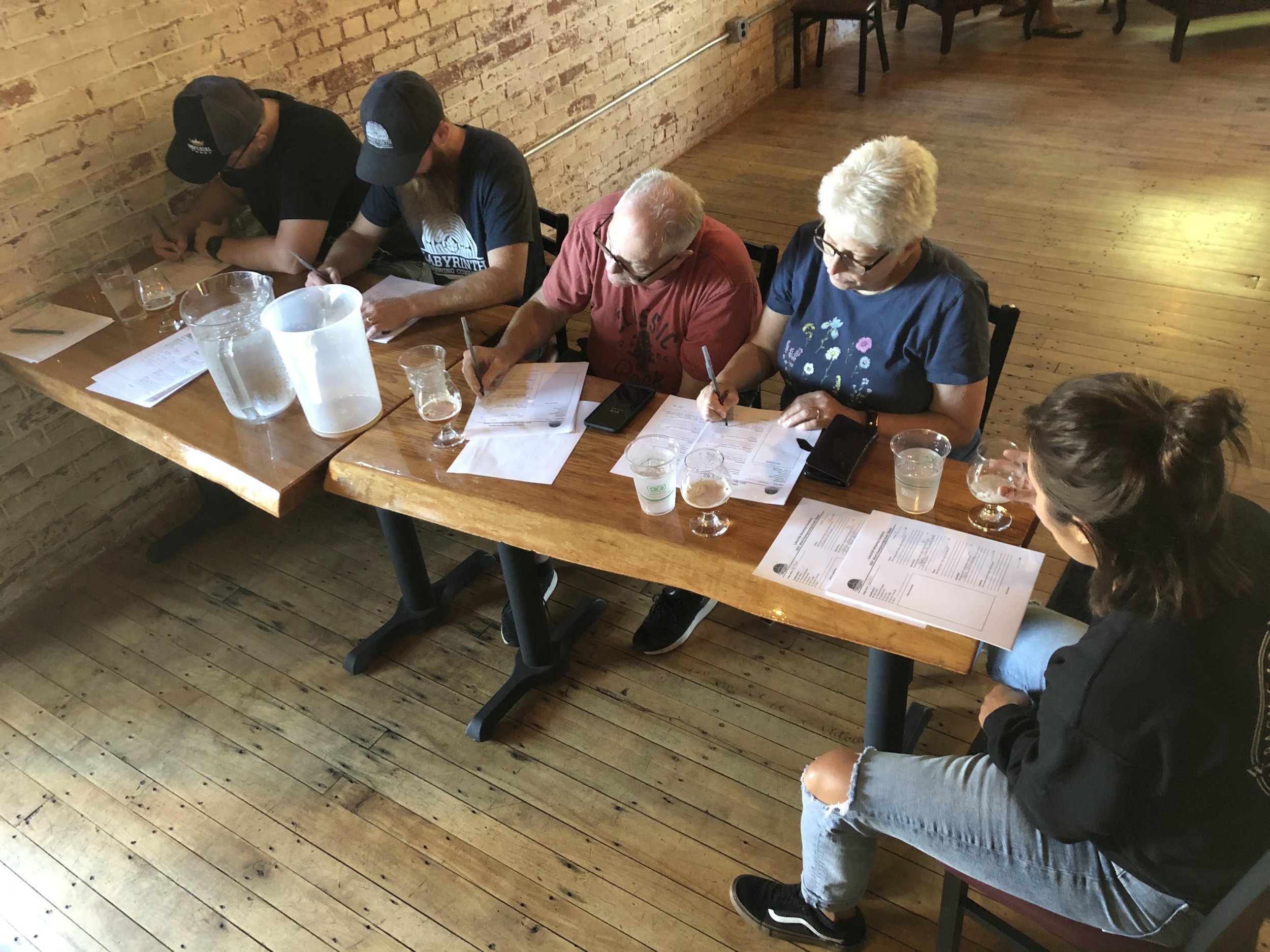 Five people seated at a wooden table examining documents, with drinks, pitchers, and a pitcher on the table in a room with brick walls and wooden floors.