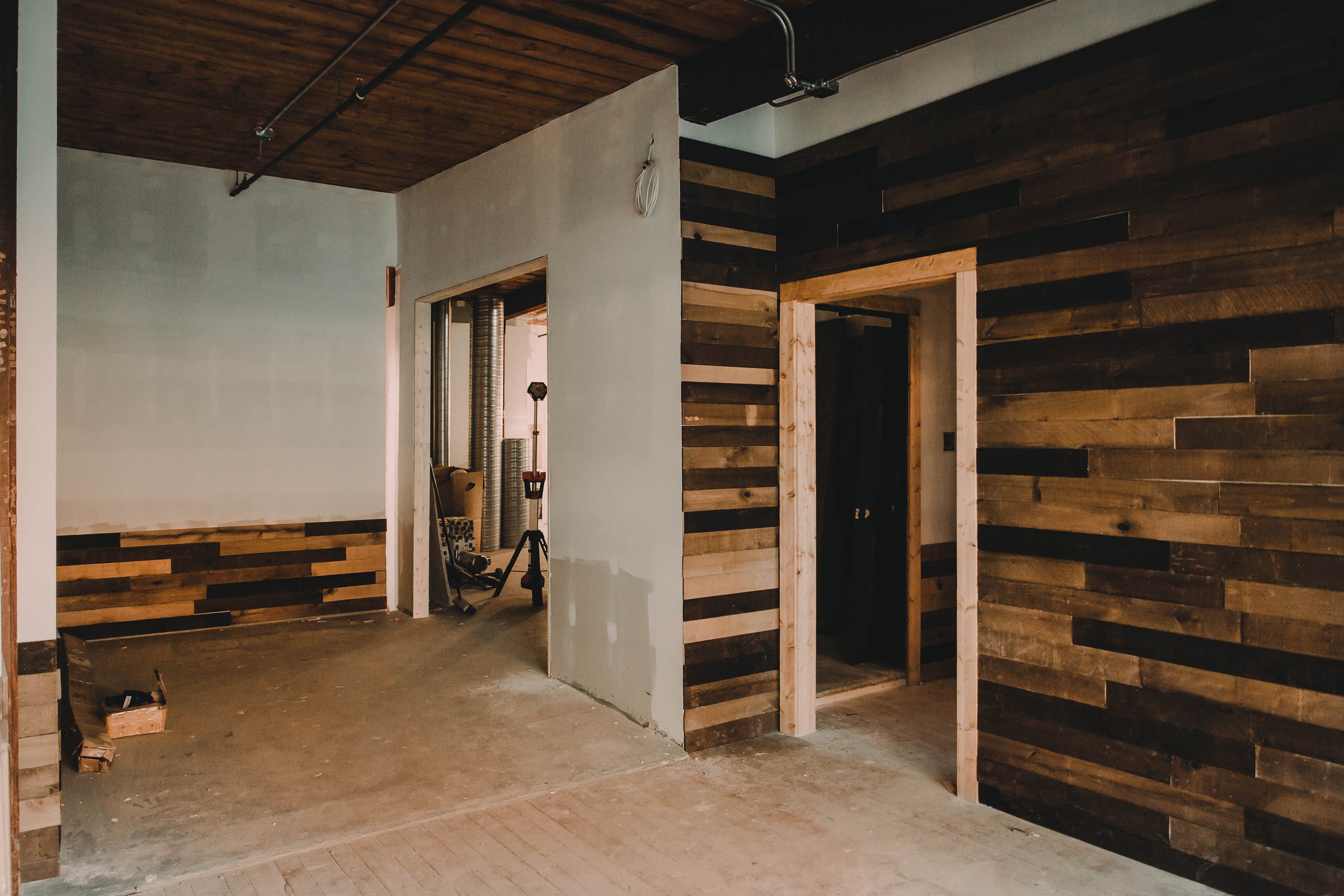Interior of a house under construction with wooden wall panels, a partially finished doorway, and construction tools visible in the background.