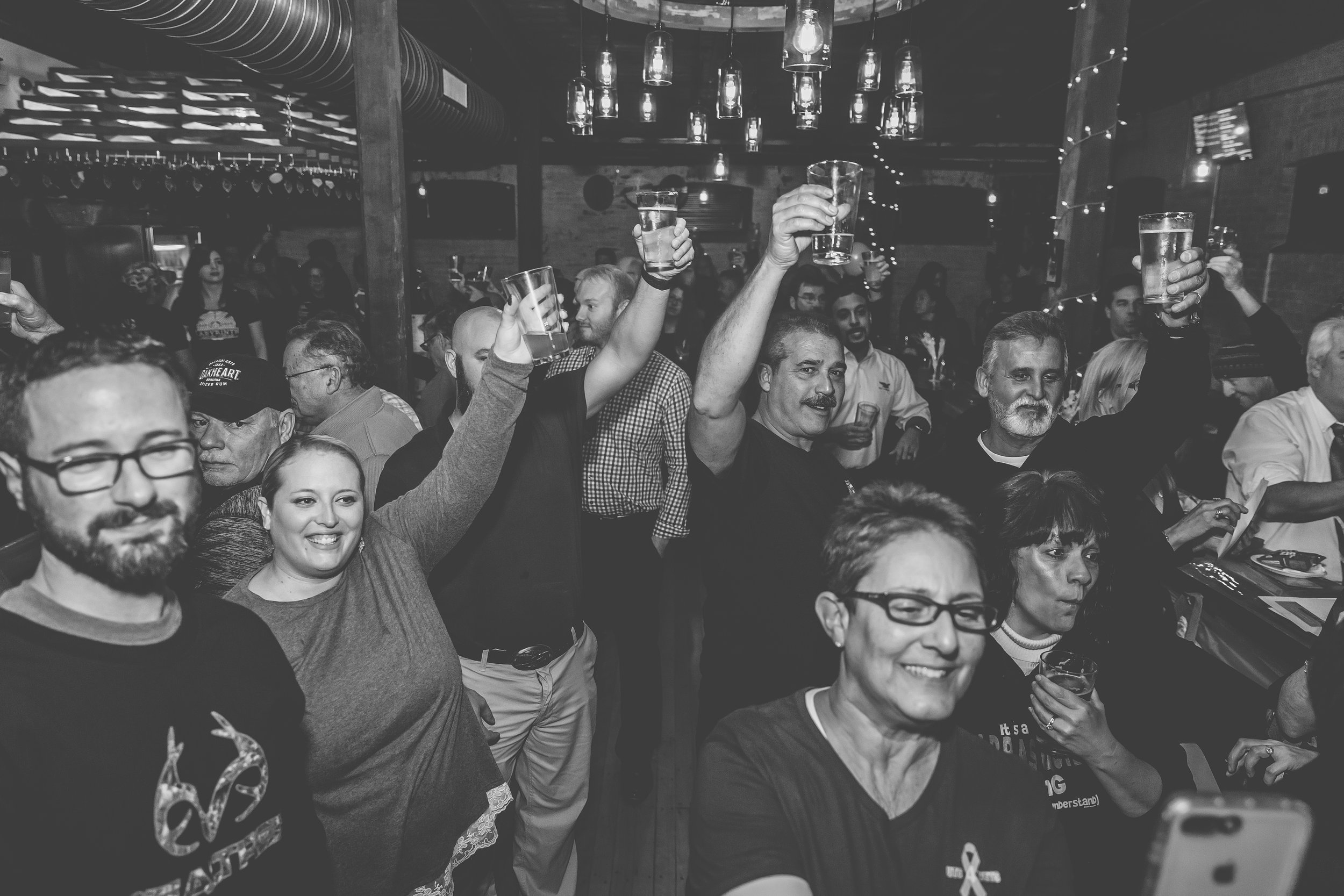A group of people raising glasses in a toast at a lively indoor celebration or concert. Black and white photo showing smiling faces and a festive atmosphere.