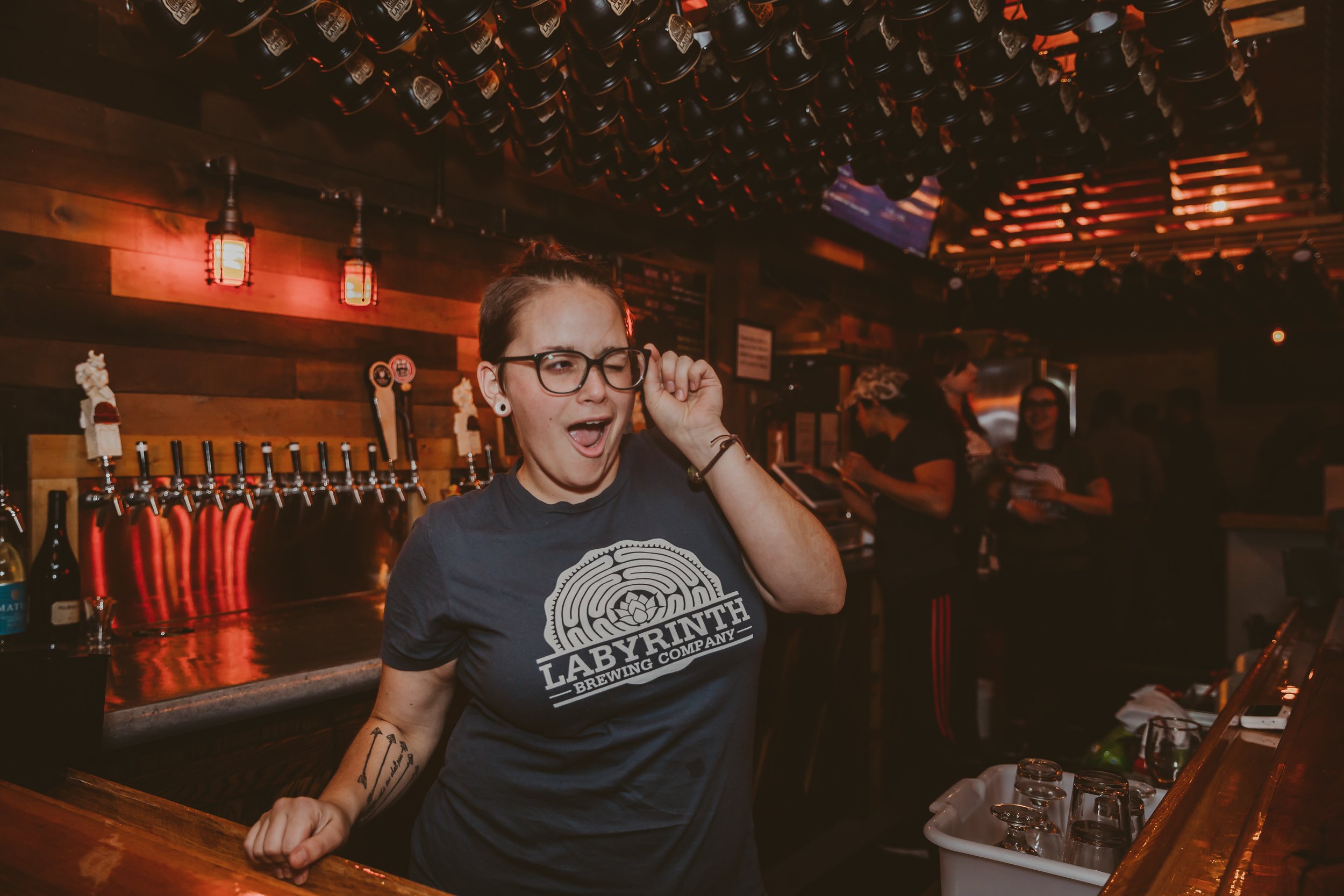 A bartender with glasses and tattoos, wearing a Labyrinth Brewing Company t-shirt, is behind the bar with a playful facial expression in a dimly lit bar. Several drink taps and glasses are visible, with other bartenders and customers in the backgroun