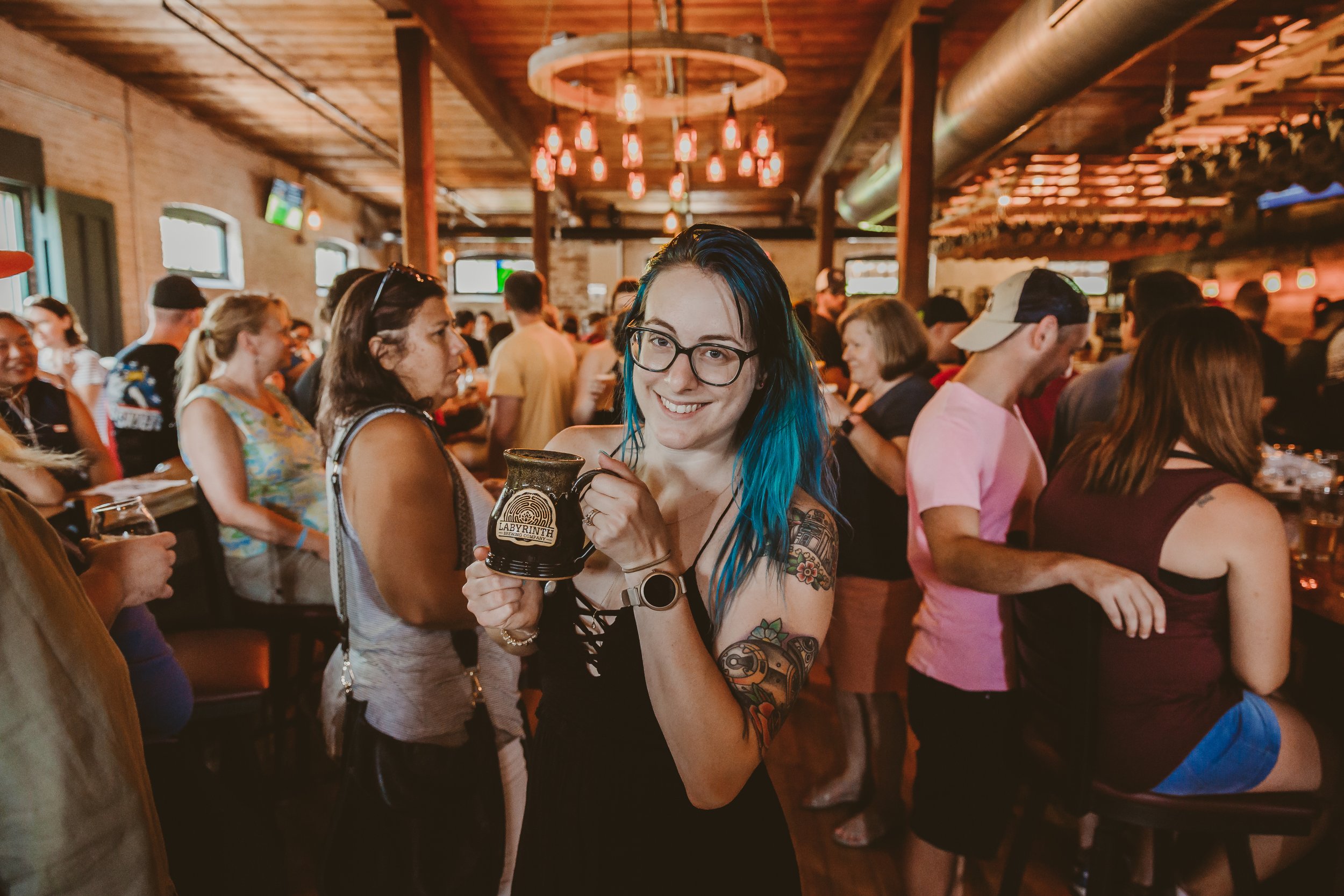 A smiling woman with blue hair and glasses holding a black mug in a busy bar or pub with many other people socializing and drinking.