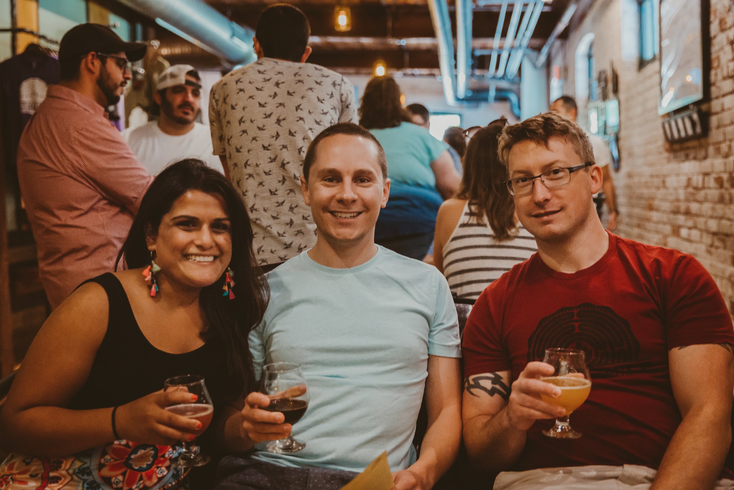 Three friends sitting at a table in a lively bar, holding drinks and smiling at the camera.