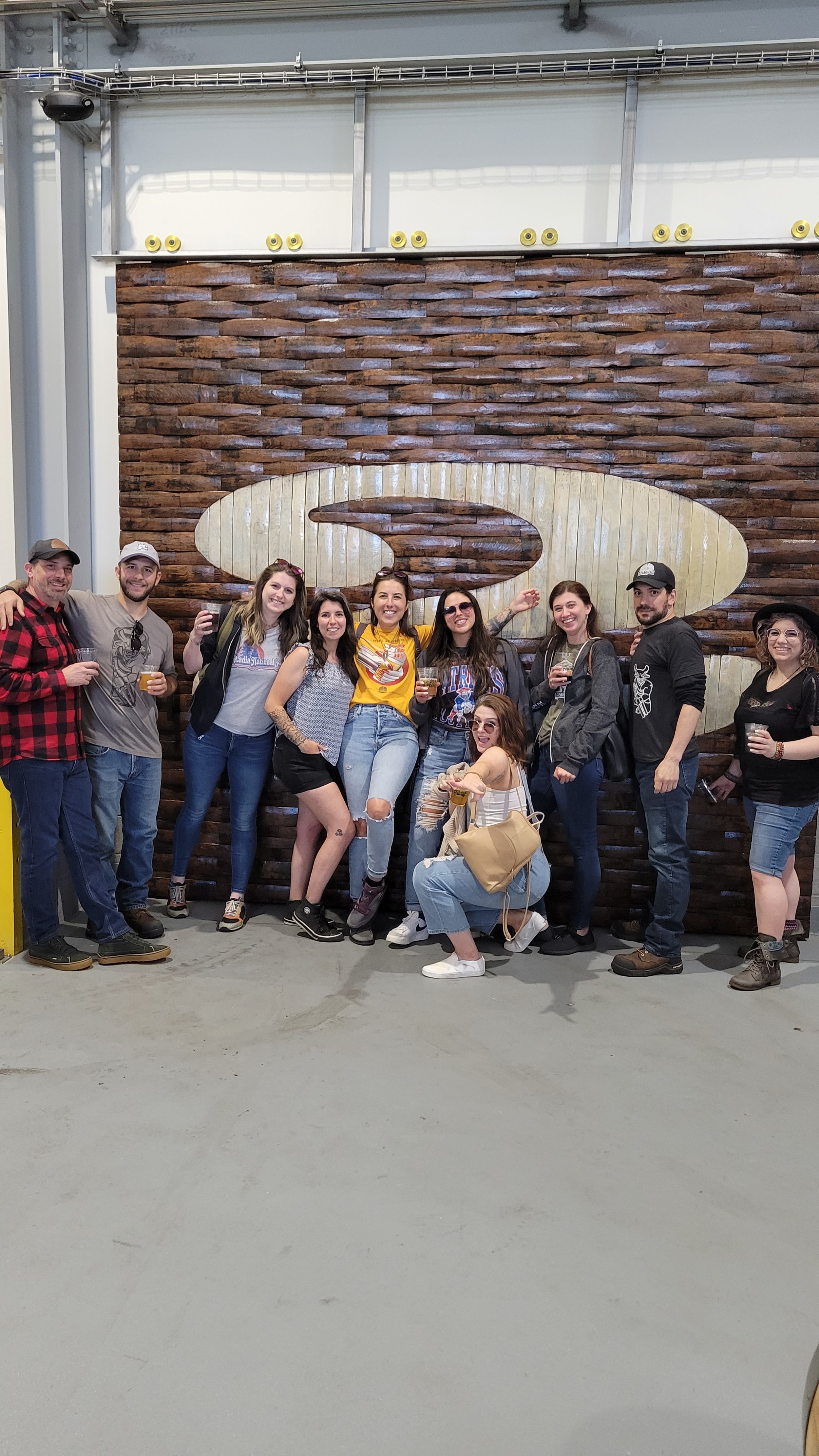 Group of ten people posing and celebrating in front of a wooden wall with a large logo, some holding drinks, in an indoor setting.