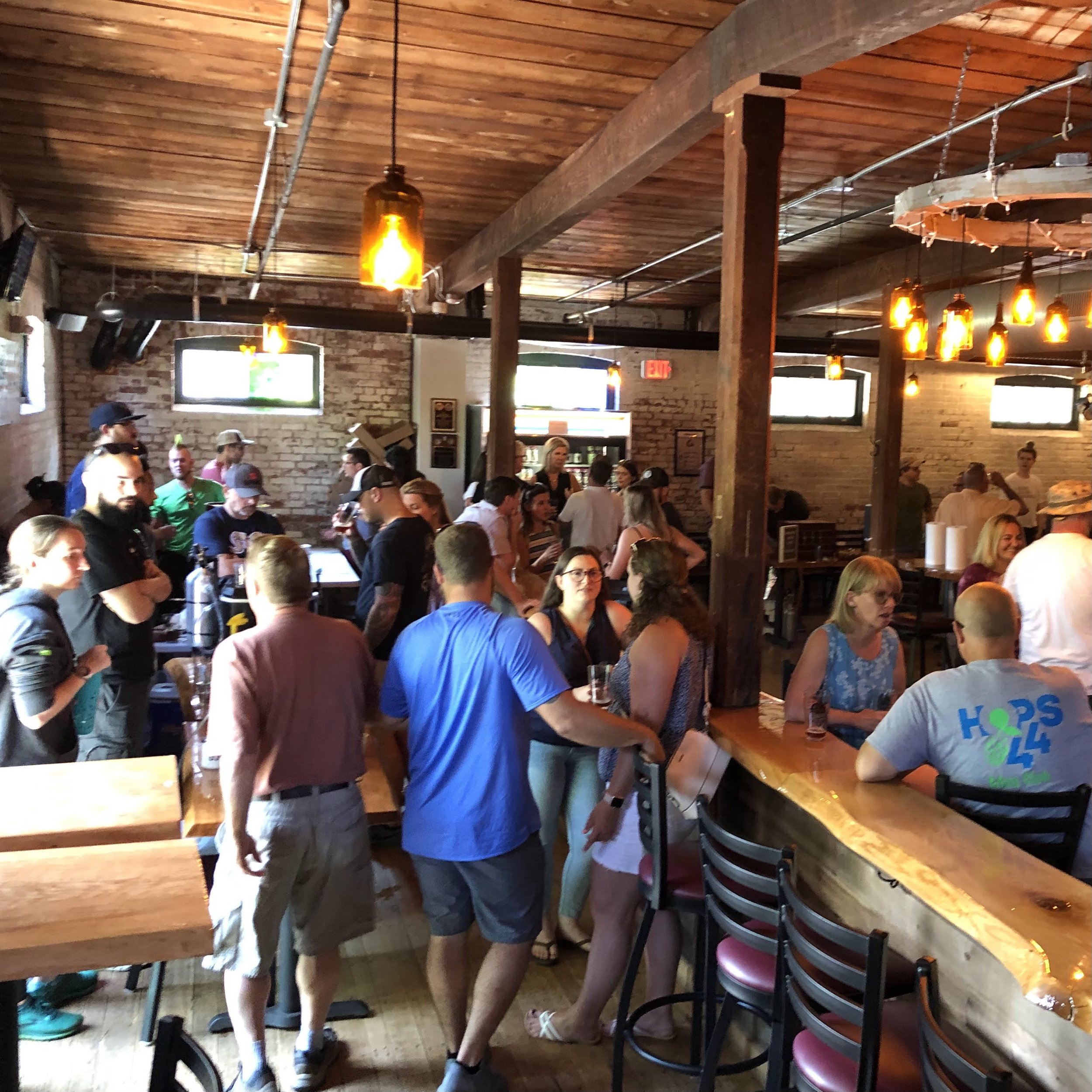 Interior of a busy bar or restaurant with people socializing and standing in line, wooden ceiling and brick walls, hanging pendant lights.