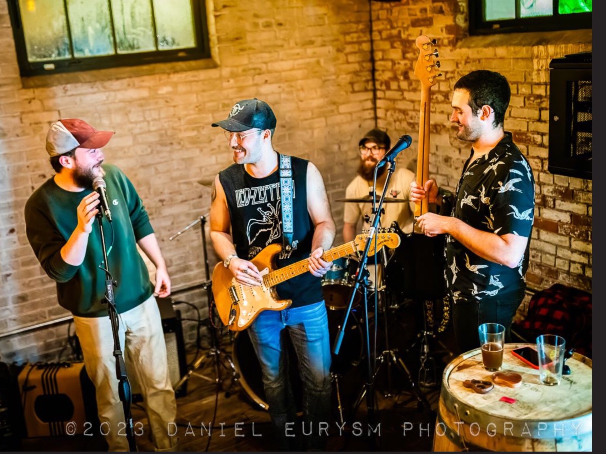 Four musicians performing in a brick-walled venue, with one person singing into a microphone, another playing an electric guitar, a third holding a bass guitar, and a drummer in the background. There are glasses and small objects on a table in the fo