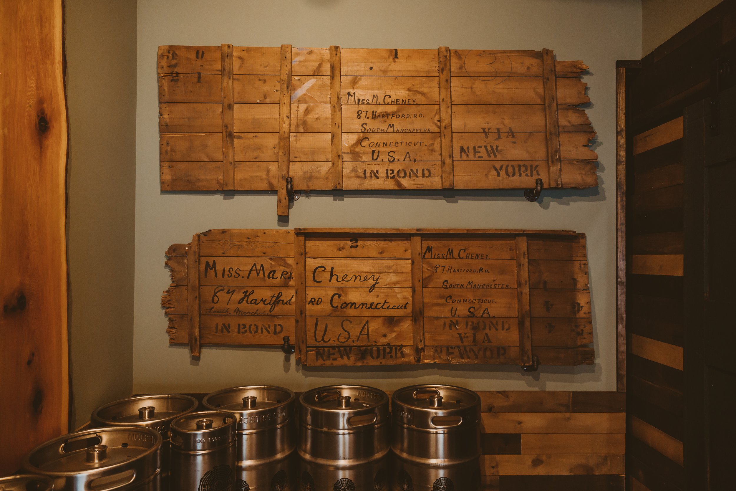 Wall with two wooden crates resembling vintage suitcase labels, mentioning Miss M. Cheney and Miss M. Mary Cheney from Connecticut to New York, along with metal beer kegs at the bottom of the wall.