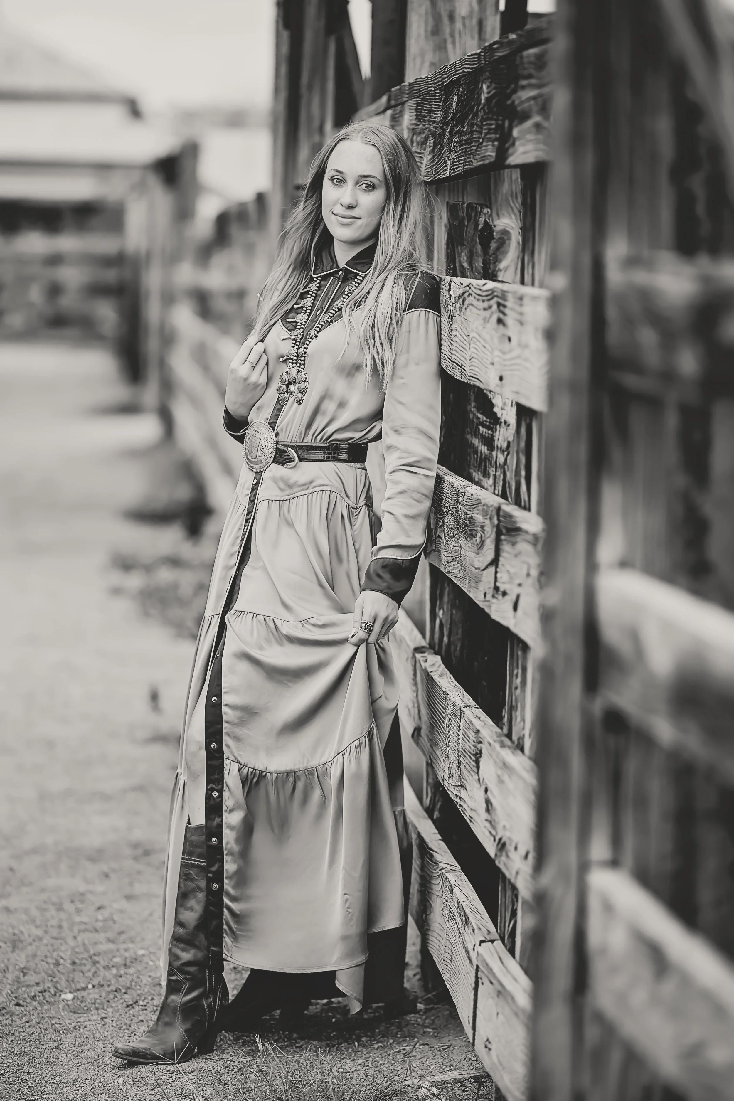 Black and white photo of a woman standing beside a wooden fence, looking at the camera, with a casual western style dress and boots.