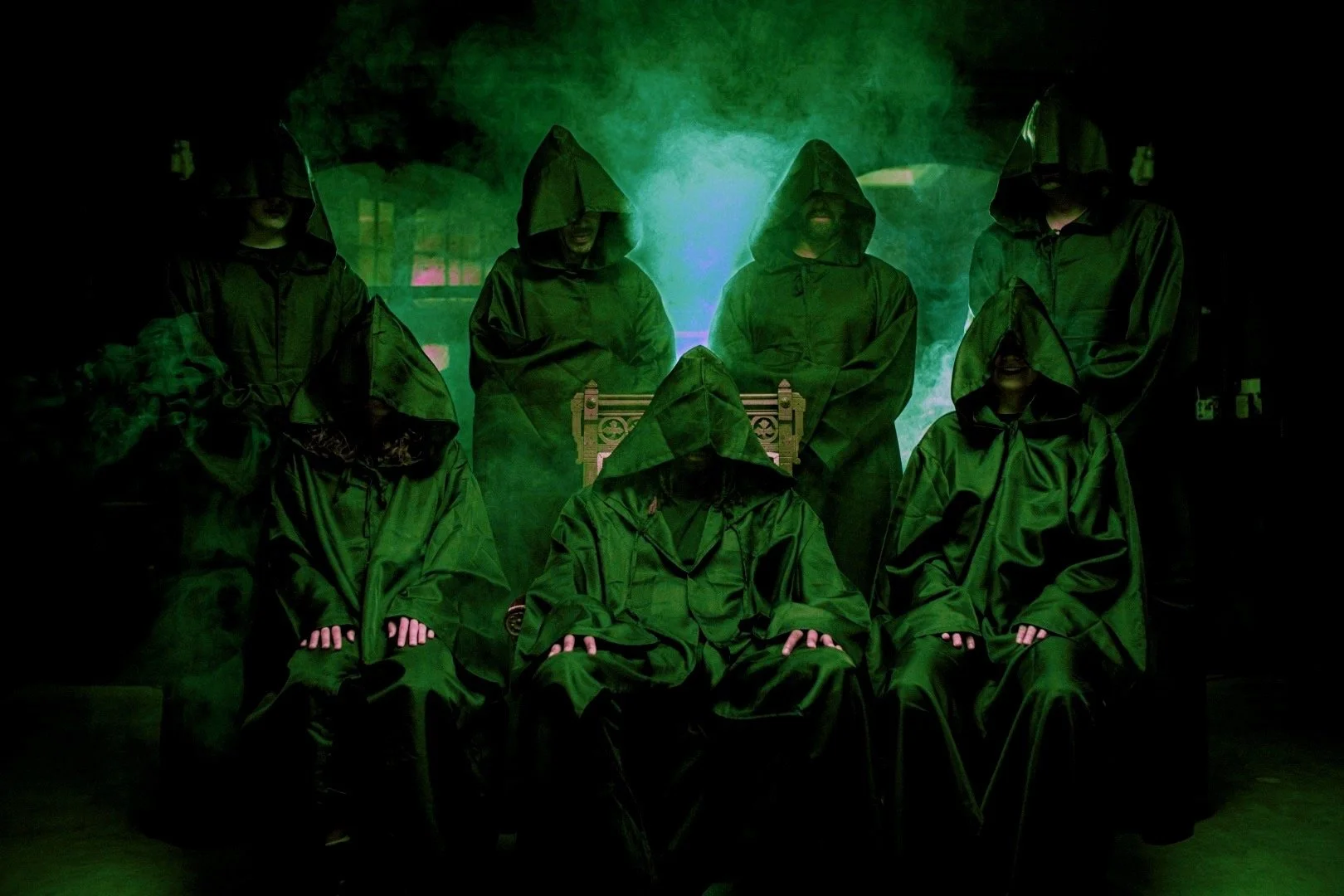 Group of people dressed in green hooded robes, some with hoods covering their faces, standing and sitting in a dimly lit room with green smoke and a glowing green light in the background. One person is seated in a chair in the center.