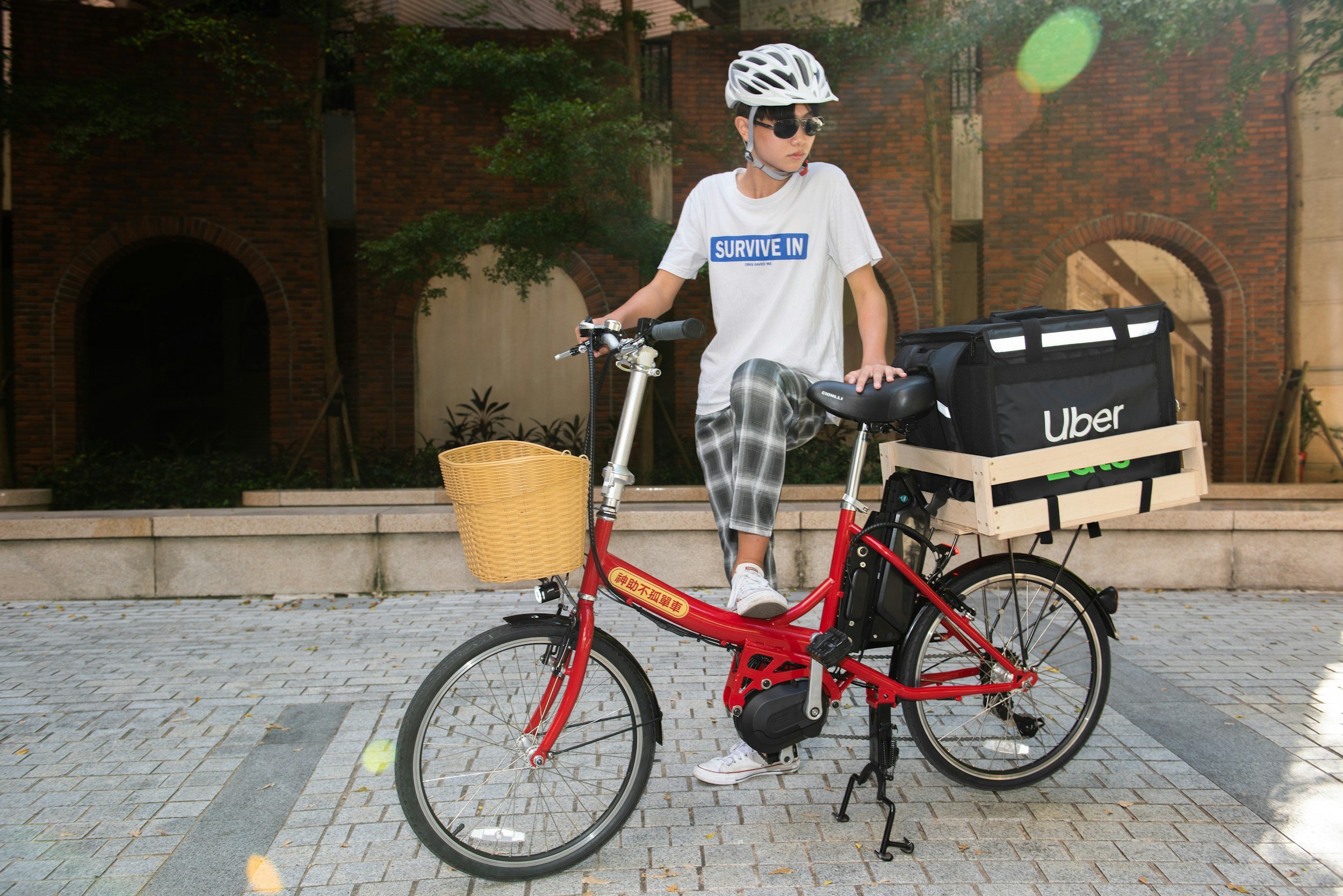 A person wearing sunglasses, a white helmet, a white t-shirt with the words 'SURVIVE IN,' and plaid pants is standing next to a red electric bike with a front basket and a rear box with Uber branding. They are outdoors on a paved area near brick buildings and greenery.