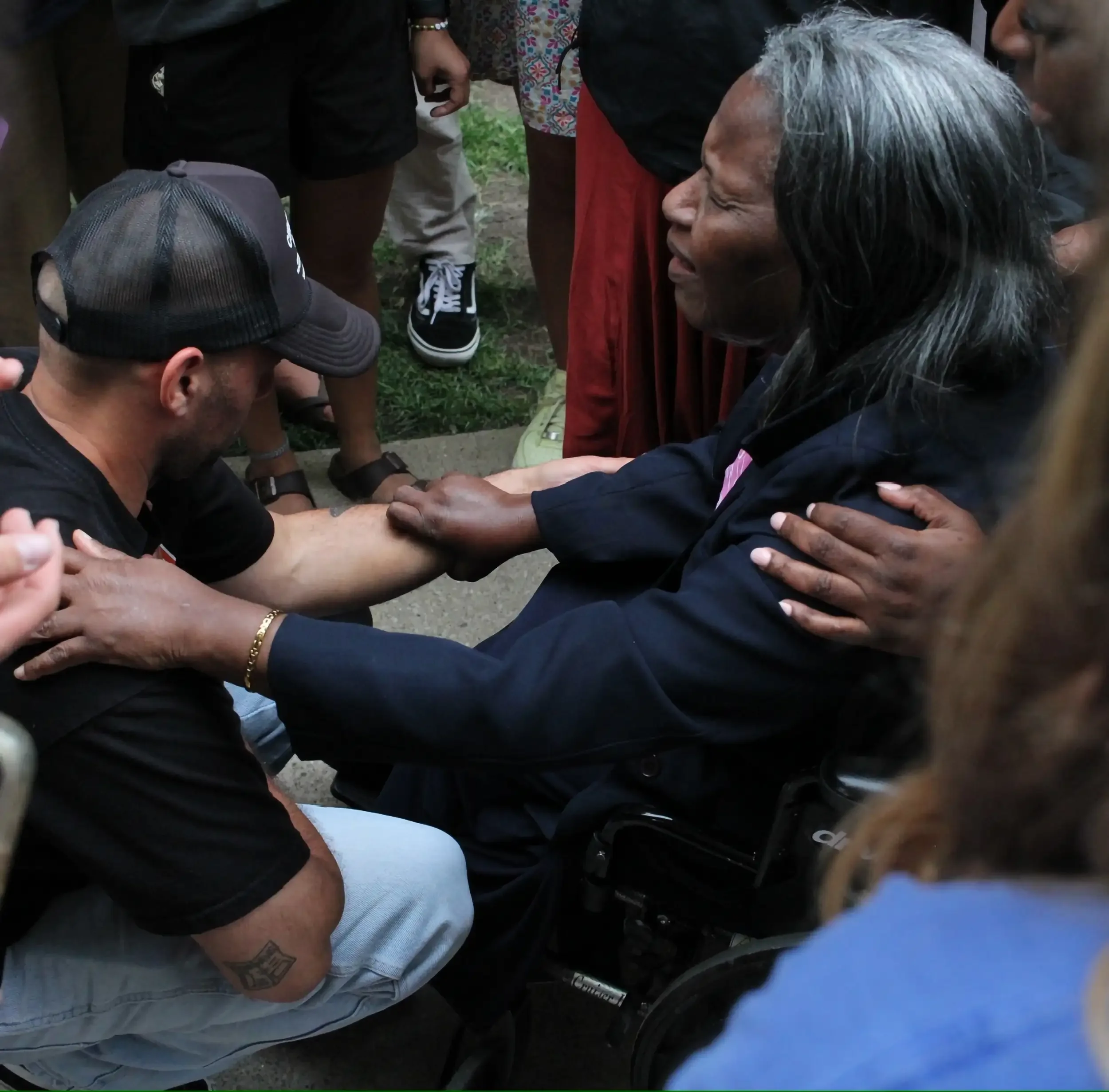 A touching moment where a woman in a wheelchair is holding hands with a man kneeling in front of her, surrounded by several people in an outdoor setting.