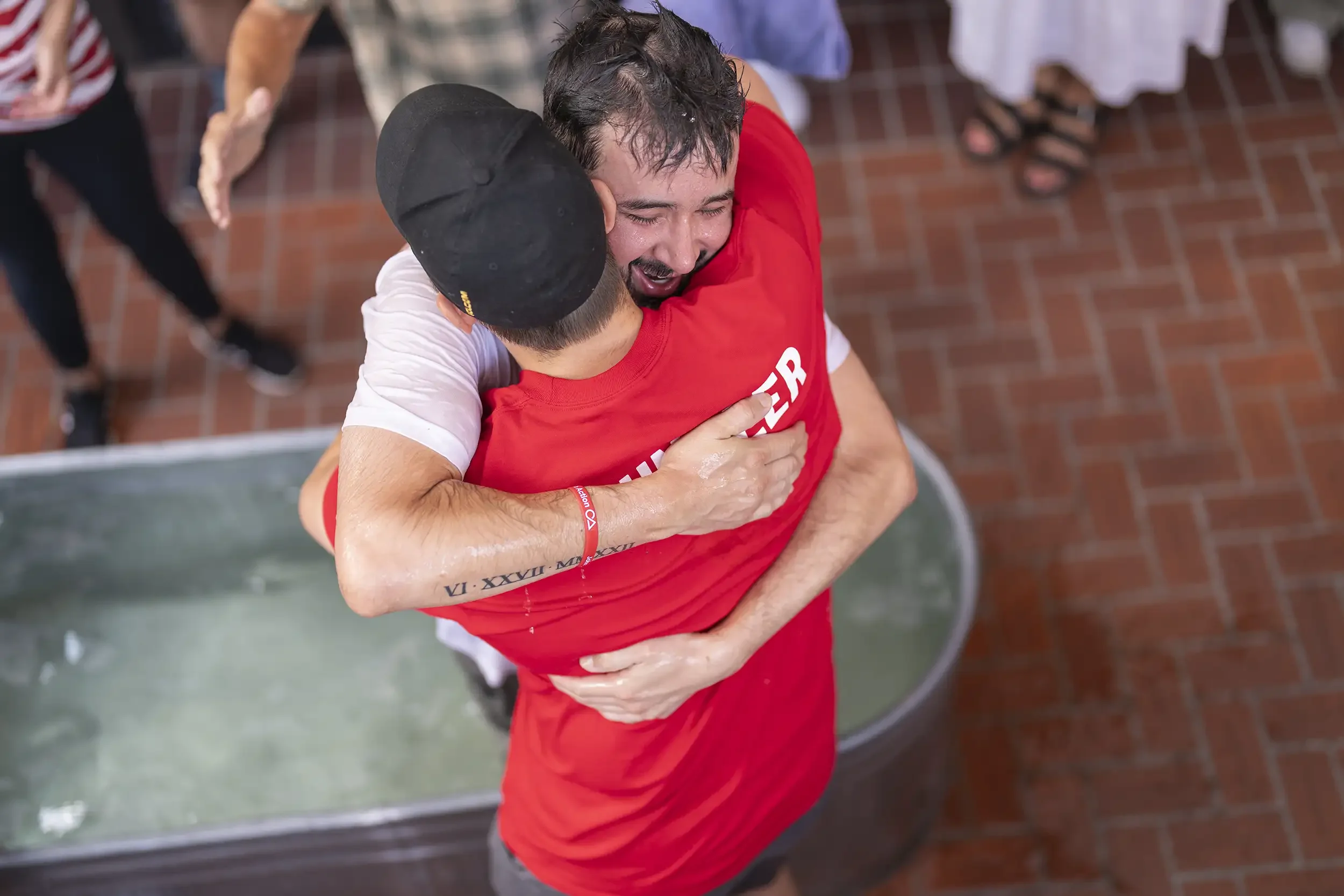 Two men are hugging each other in celebration, one wearing a white shirt and the other a red shirt, with water on their skin. They are smiling with joy. The background shows a brick floor and a metal baptismal font filled with water.