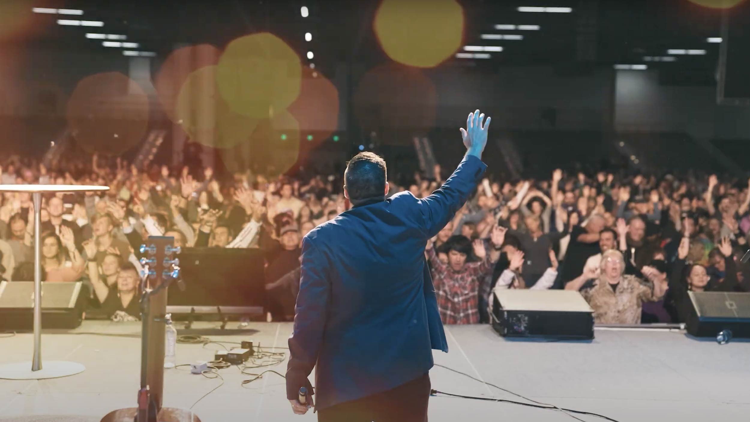 A speaker on a stage waving to a large audience in a dark auditorium.