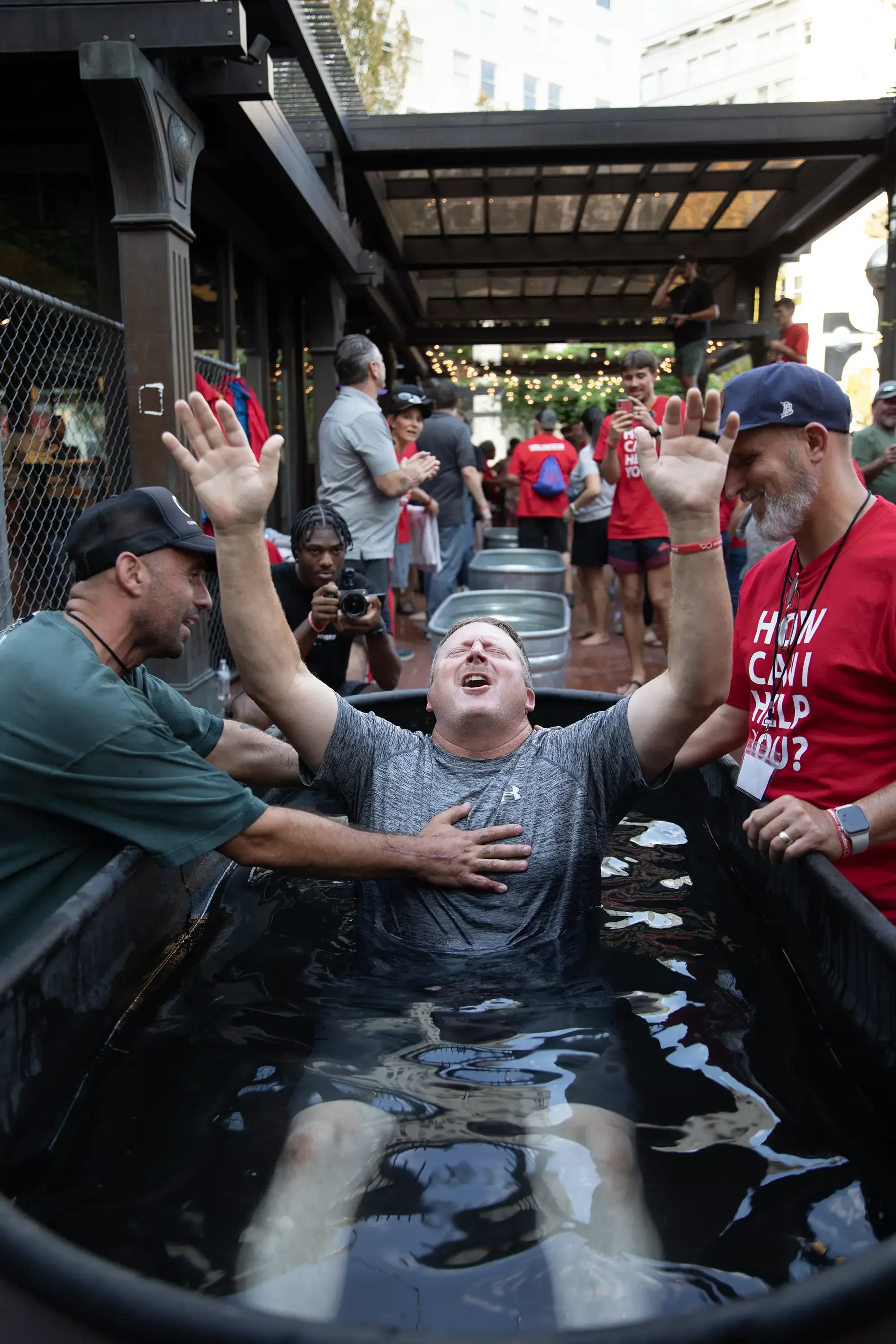 A man is being baptized in a water container, surrounded by smiling people, with one hand on his chest and the other raised in the air, during an outdoor church service or event.
