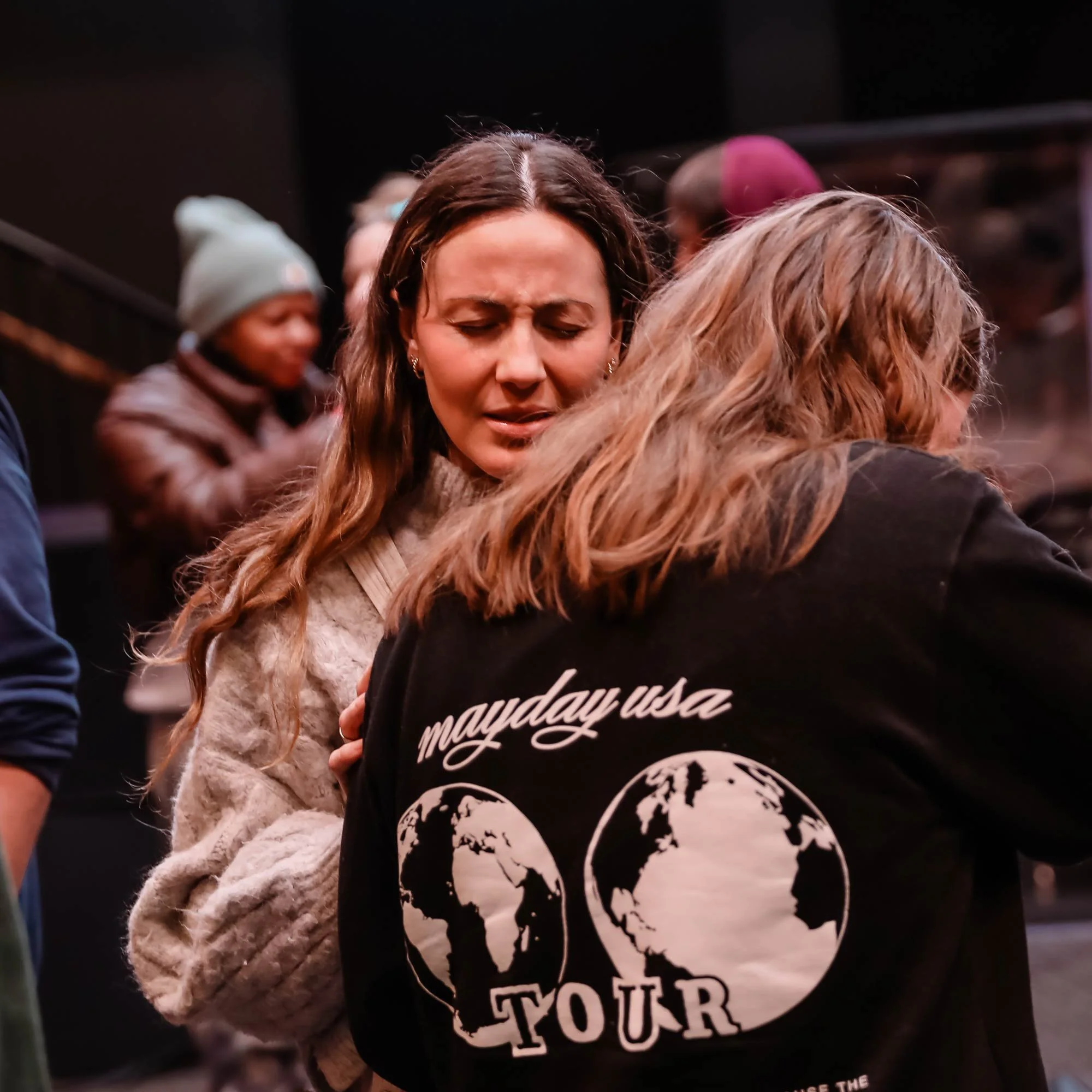 A woman with long brown hair, wearing a beige sweater, appears emotional with her eyes closed and a distressed expression. She is hugging or leaning against another woman with shoulder-length brown hair, wearing a black jacket with the white text 'mayday usa TOUR' and a globe graphic on the back. In the background, other people are visible, including a person wearing a gray beanie and brown jacket.
