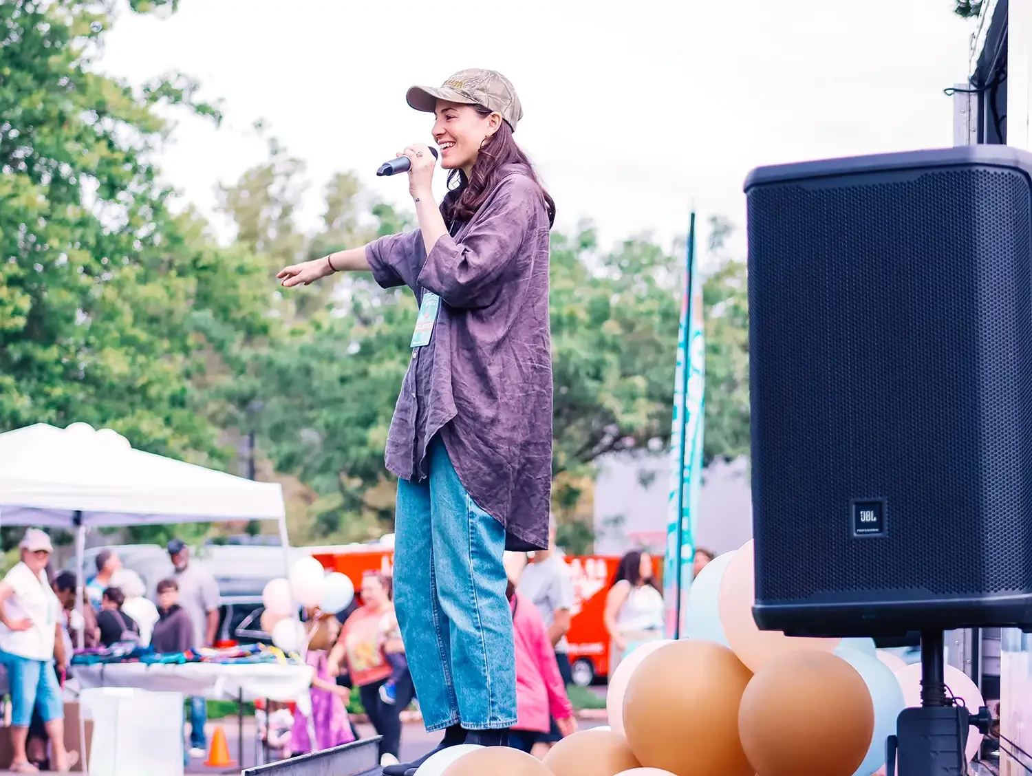 A woman wearing a cap and casual clothes is speaking or singing into a microphone at an outdoor event, with people and tents in the background.