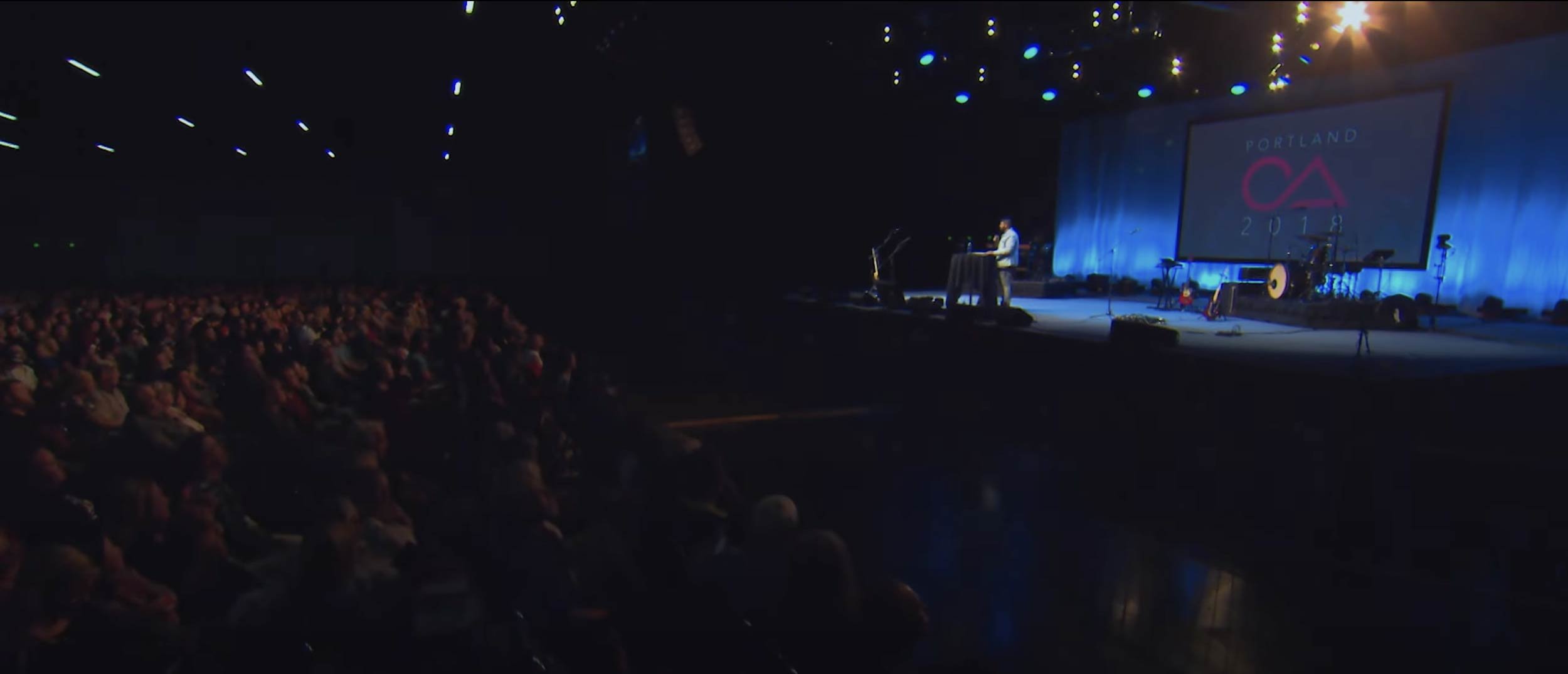 A large gathering of people sitting in an auditorium watching a stage presentation with a blue backdrop and a large screen displaying 'Portland 2018' and a pink logo.