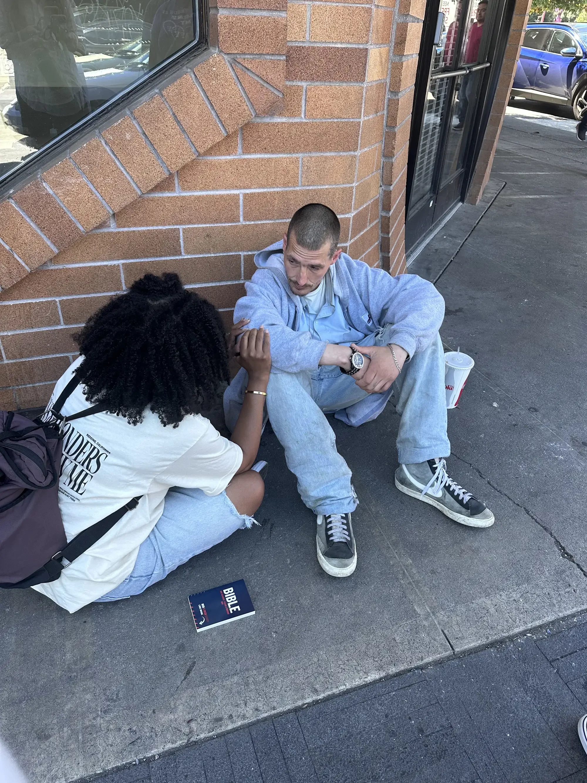 A young woman and a young man sit on the sidewalk outside a building with a brick exterior. The woman, with curly black hair, is holding the man's arm and appears to be praying or comforting him. The man, with short hair, wearing a grey hoodie, light blue shirt, and ripped jeans, looks distressed. There is a Bible placed on the ground near them and a large drink cup nearby.