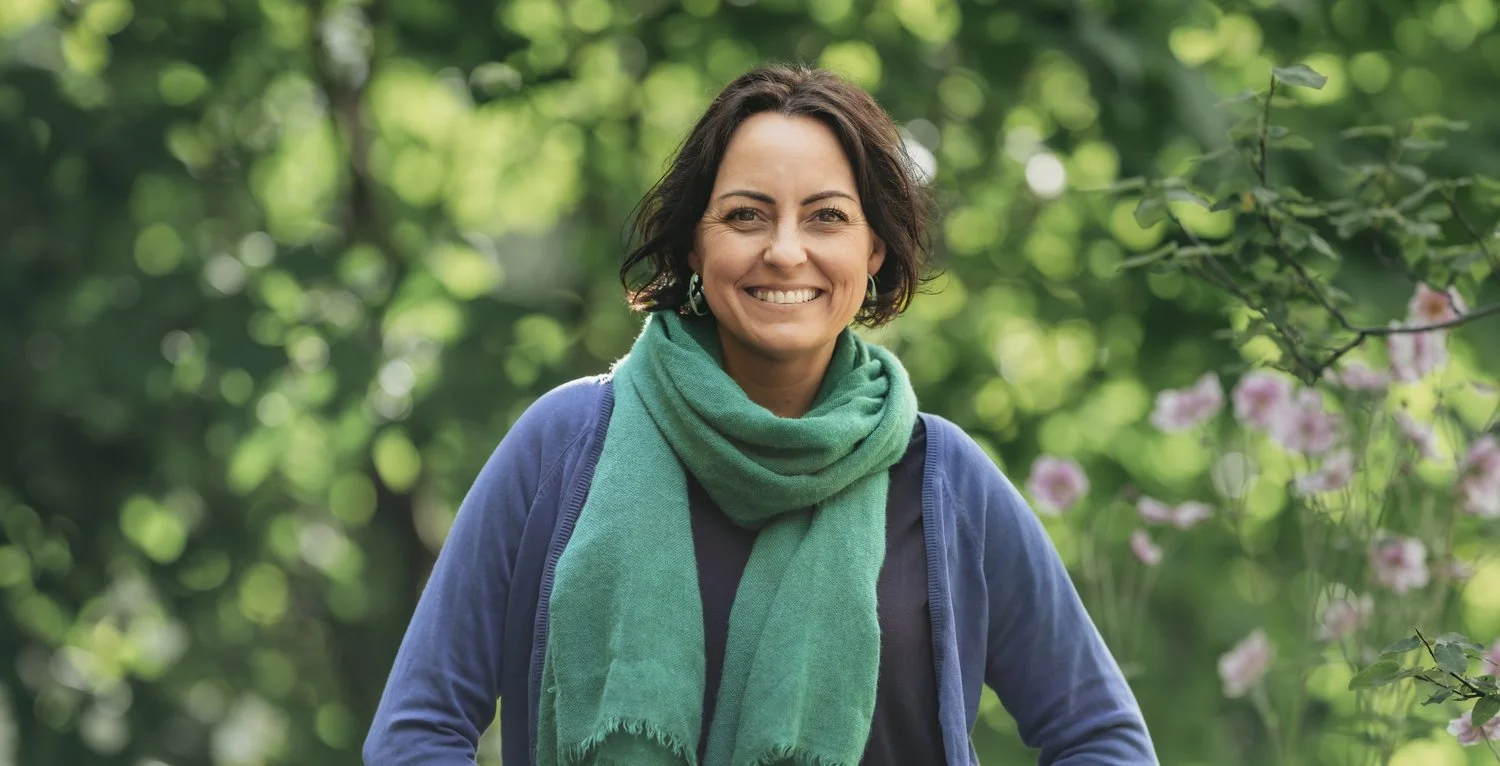Sarah Kirby of Fuse Fitness & Massage poses in a green garden, smiling at the camera. Sarah is wearing a blue cardigan and teal scarf. Her brunette hair is styled in a short bob.