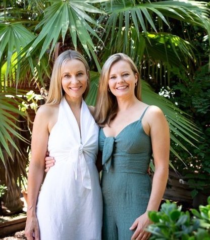 Nicole and Kelly, co-founders of Abundance Unlimited, are standing in a leafy tropical garden, smiling at the camera and wearing linen jumpsuits