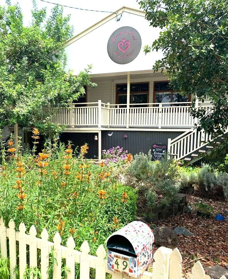 Colorful garden with orange flowers, green foliage, and a white picket fence in front of a two-story building with a balcony. The building has a circular sign with pink lettering that says 'Heart & Soul of Wellness' and a pink heart logo.