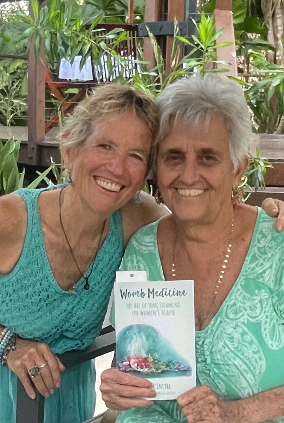 Two smiling women, with short, gray hair sitting close together outdoors, holding a book titled 'Womb Medicine' with a colorful cover. One woman is wearing a turquoise lace top and the other is in a green patterned dress. Green plants surround them.