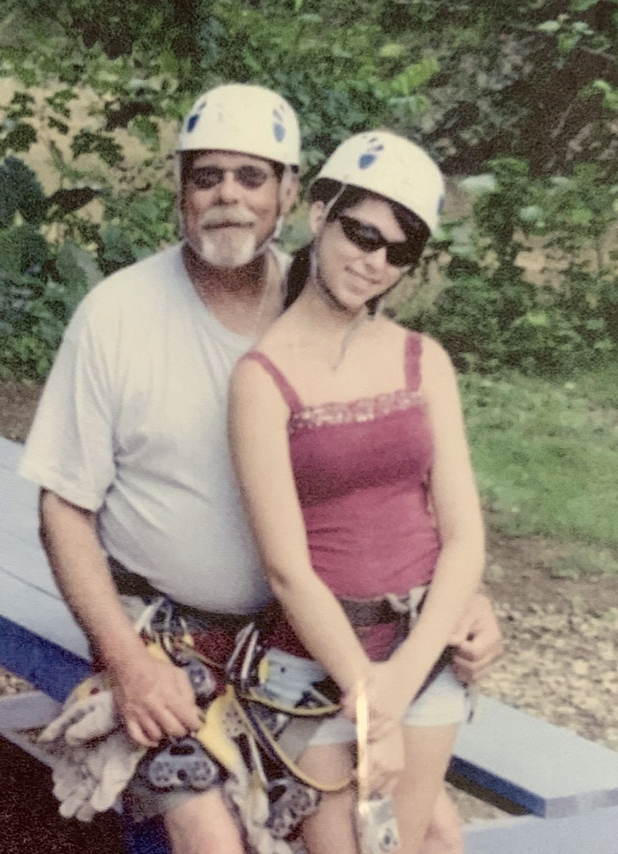 Older man and young woman wearing helmets and harnesses, sitting on a bench outdoors in a wooded area.